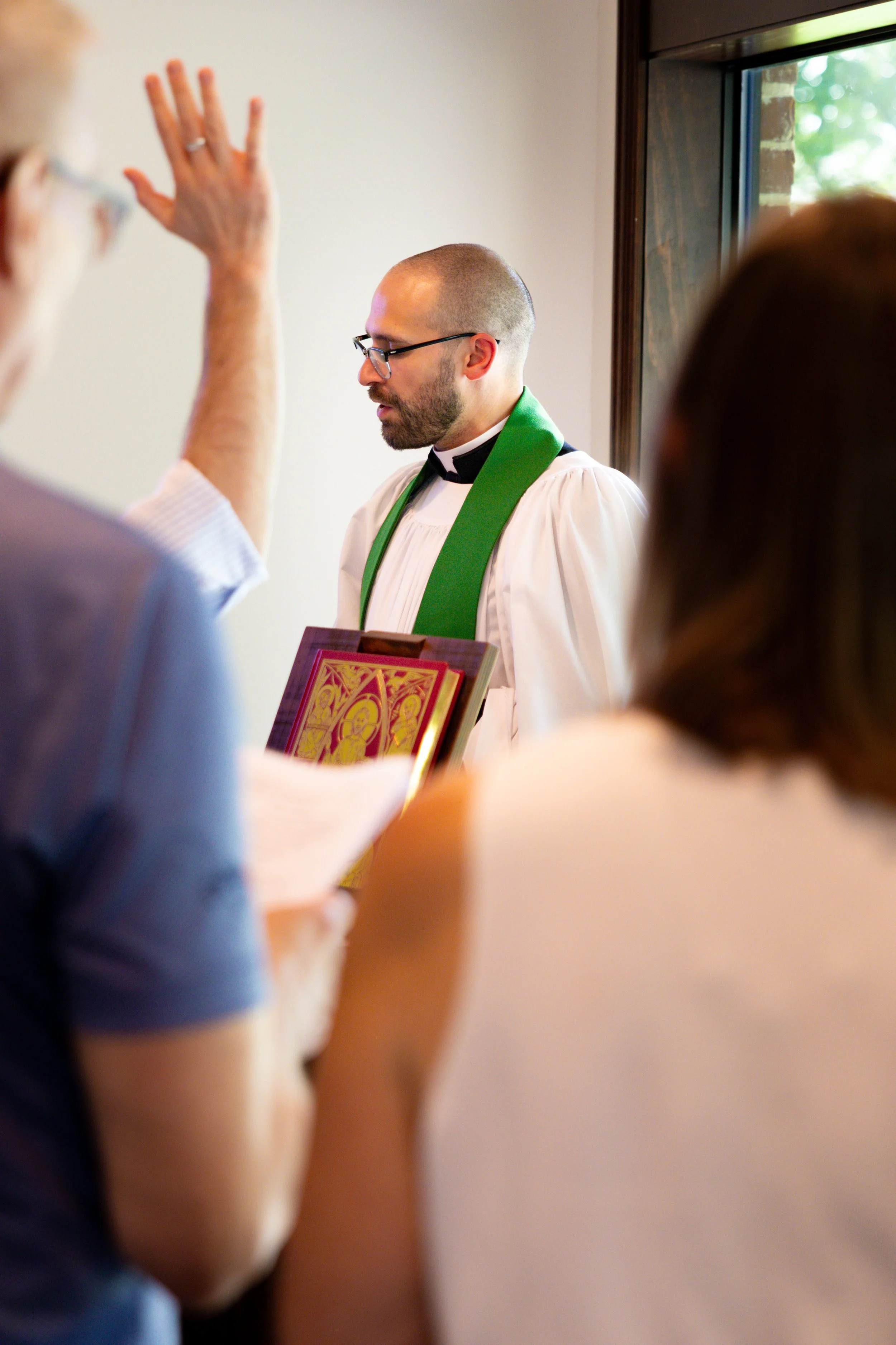 A priest with glasses and a beard, dressed in a white robe with a green stole, standing with eyes closed during a Sunday communion service.