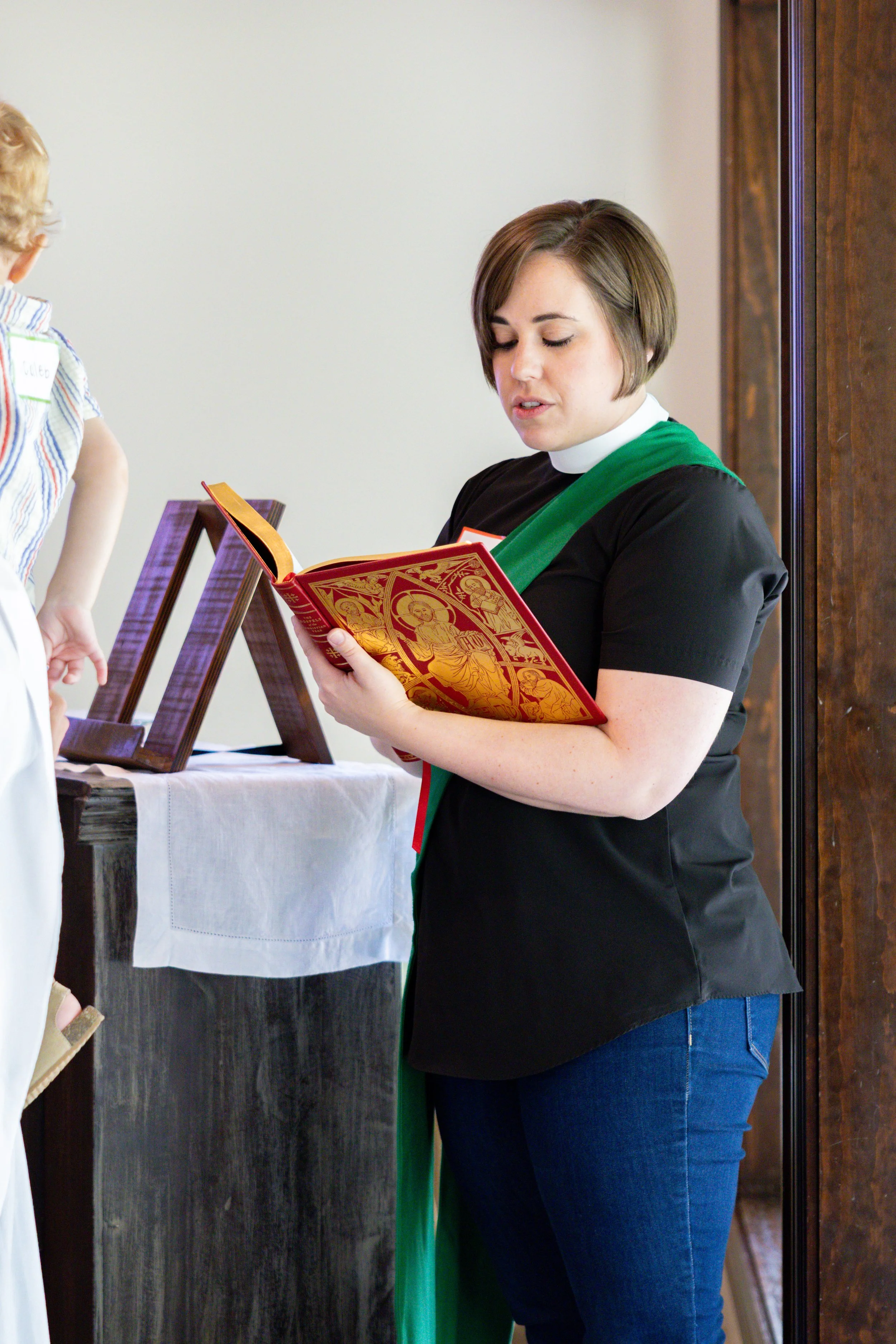 A woman in a black shirt with a green stole is reading from a red and gold religious book, standing indoors near a table with a white cloth, and a small wooden stand with religious icons.