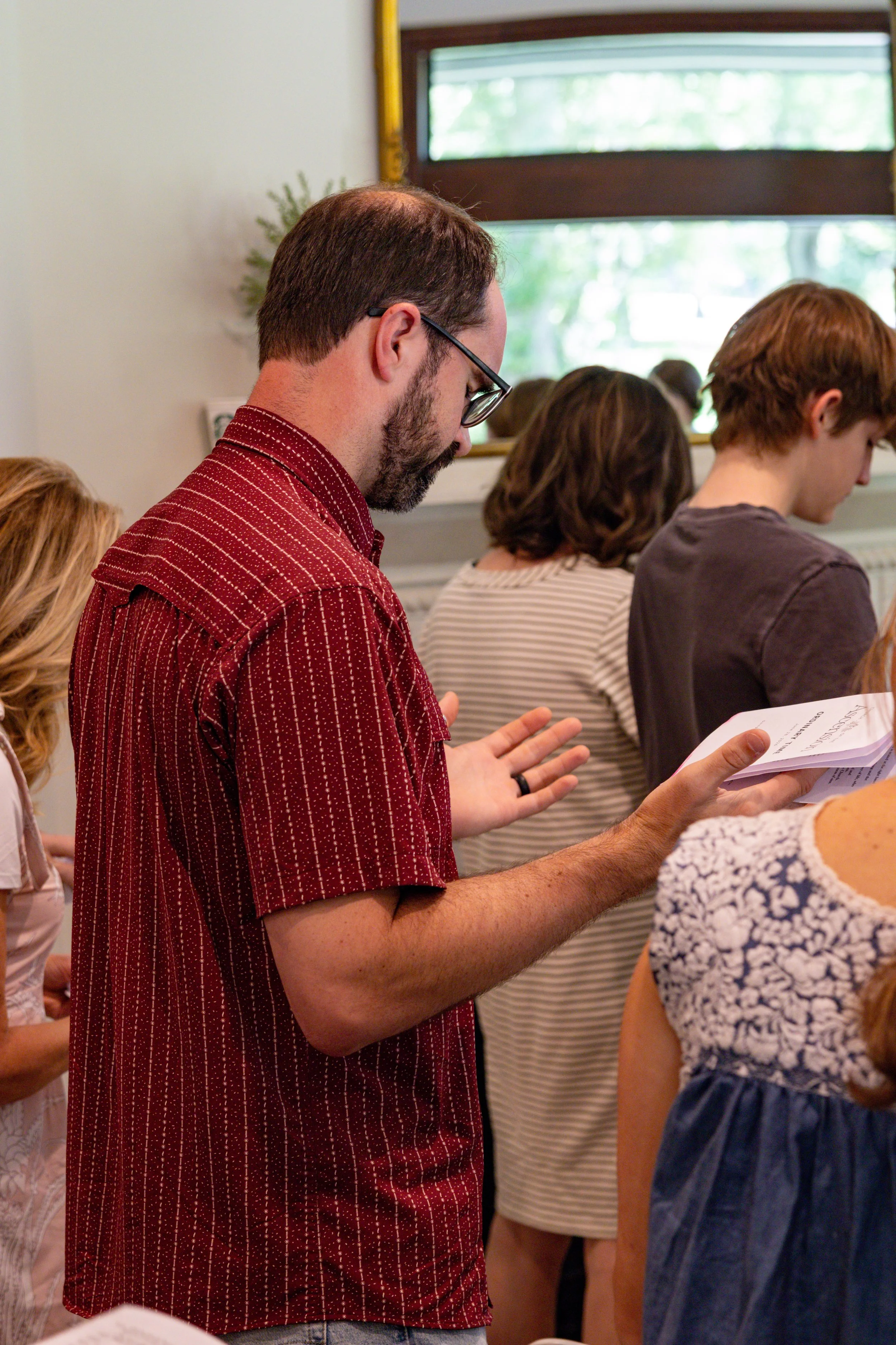 A group of people standing closely together, engaged in reading or prayer, in a room with a window and natural light.