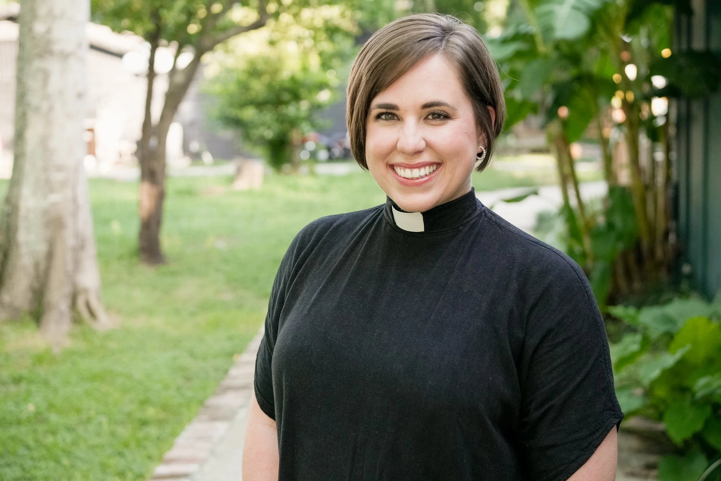 A smiling woman dressed as a priestess standing outdoors on a sidewalk with trees and greenery in the background.