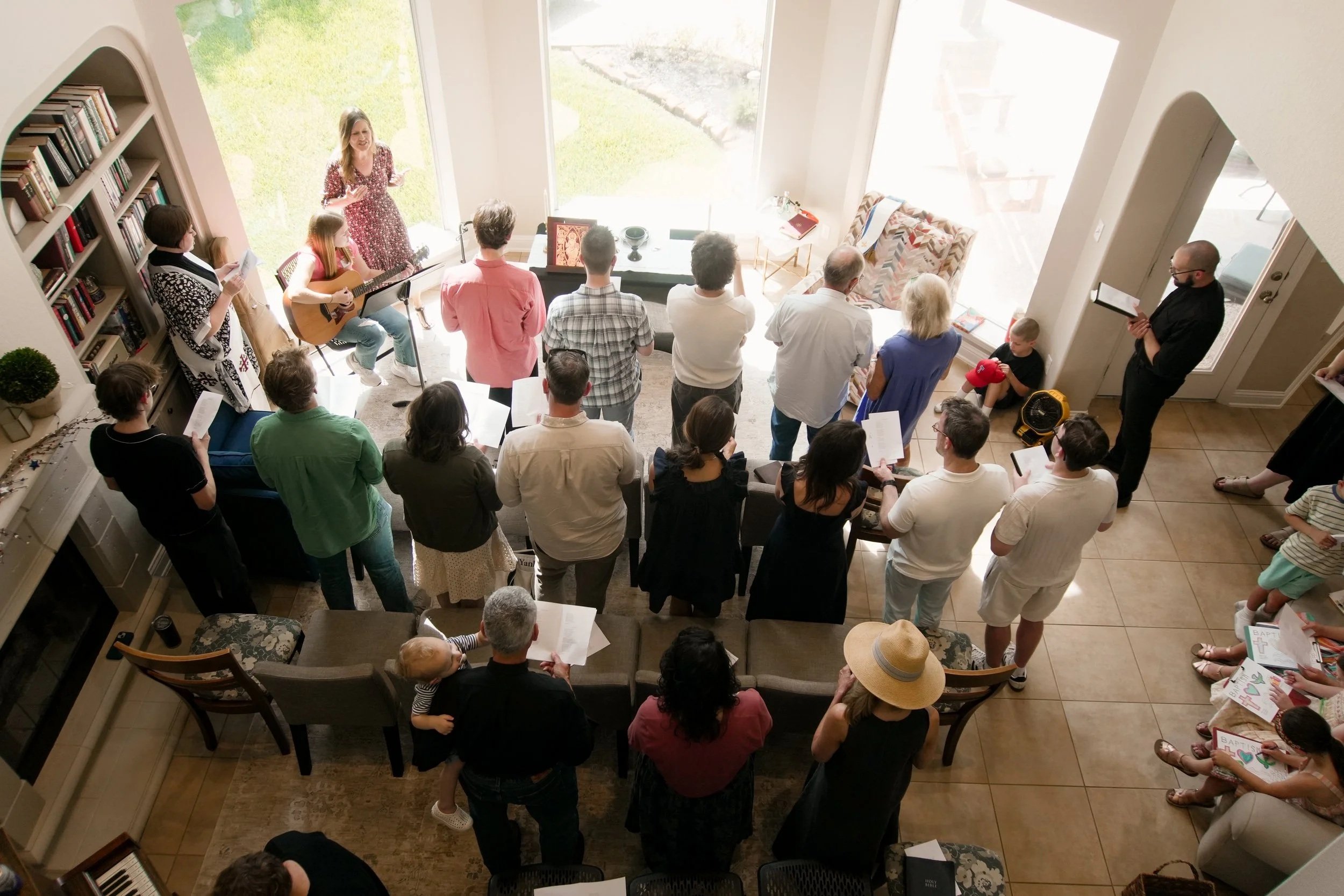 A group of people gathered indoors for worship. A woman is playing guitar and singing at the front, while others stand and hold song sheets. The room is bright with large windows, bookshelves, and cozy furniture.