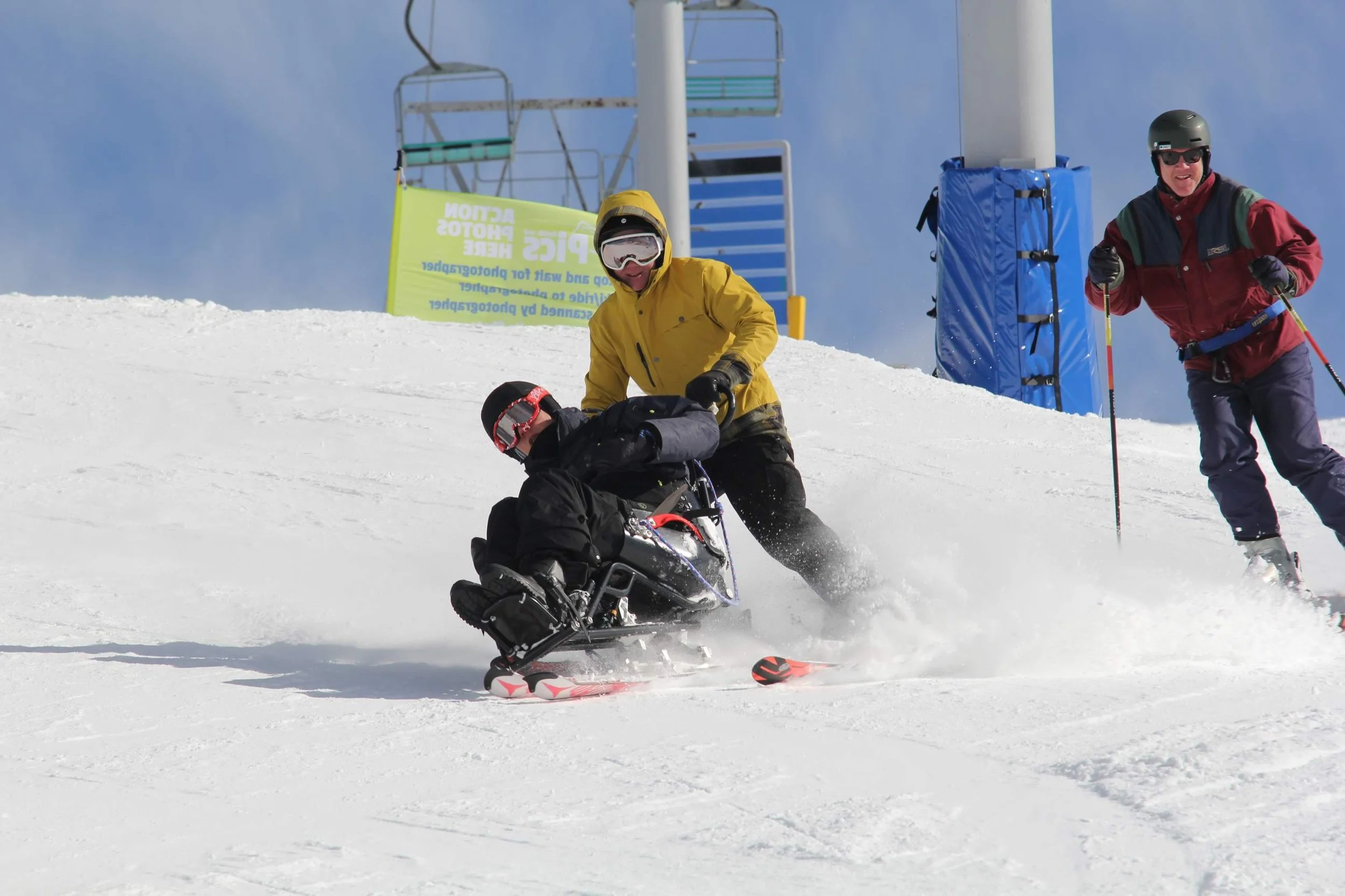 Two skiers and a person on a snowmobile on a snowy mountain slope, with ski lift structures and signage in the background.