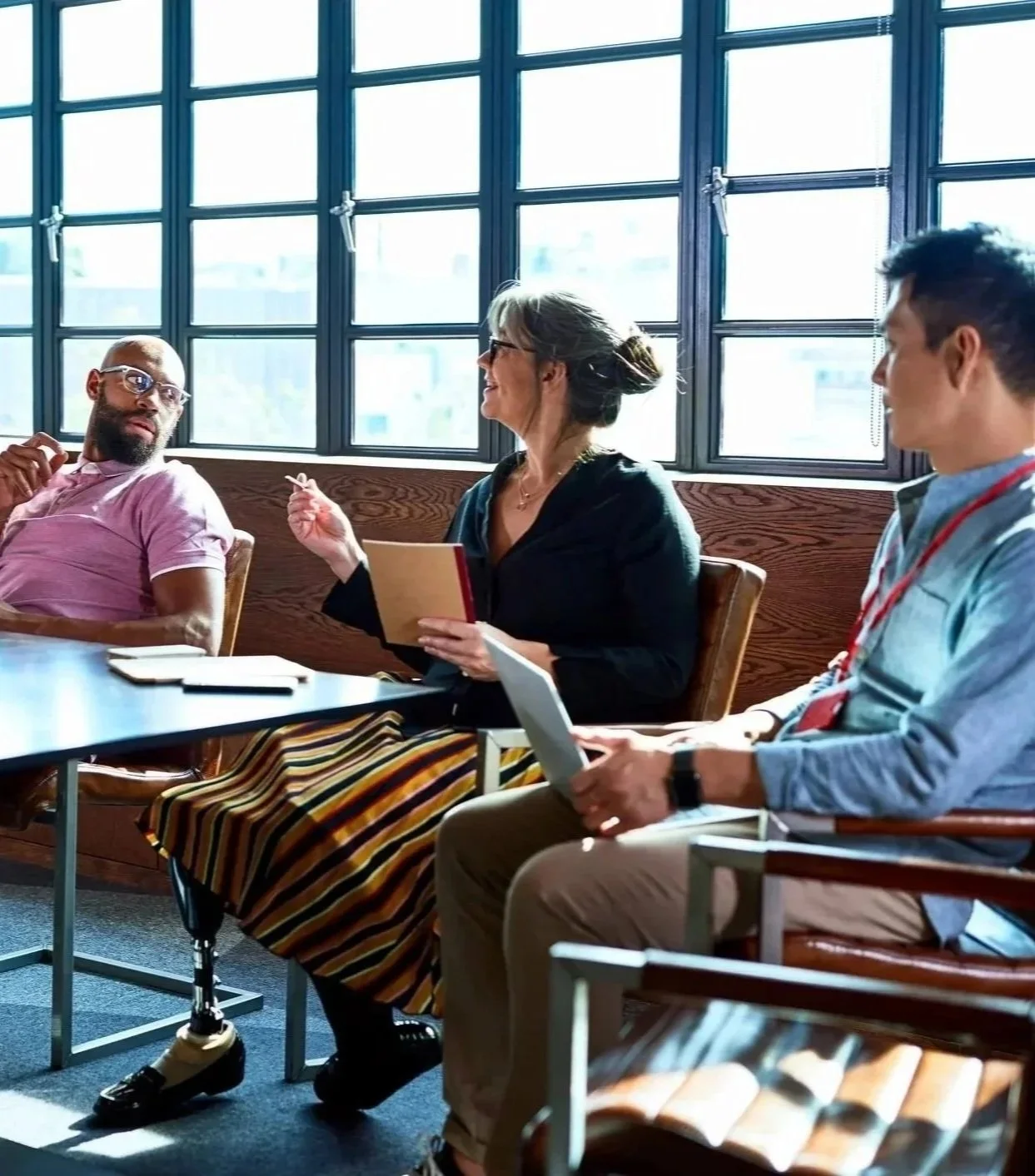 Three people engaging in a discussion in a bright conference room with large windows, including a woman with gray hair and glasses holding a notebook, a man in a pink shirt with glasses, and a young man with dark hair and a blue shirt, all seated around a table.