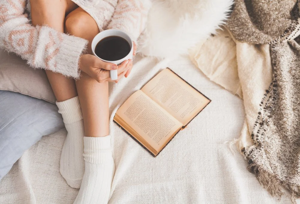 Person sitting on a cozy bed, holding a mug of coffee or tea, with a book open on the bed and blankets around.