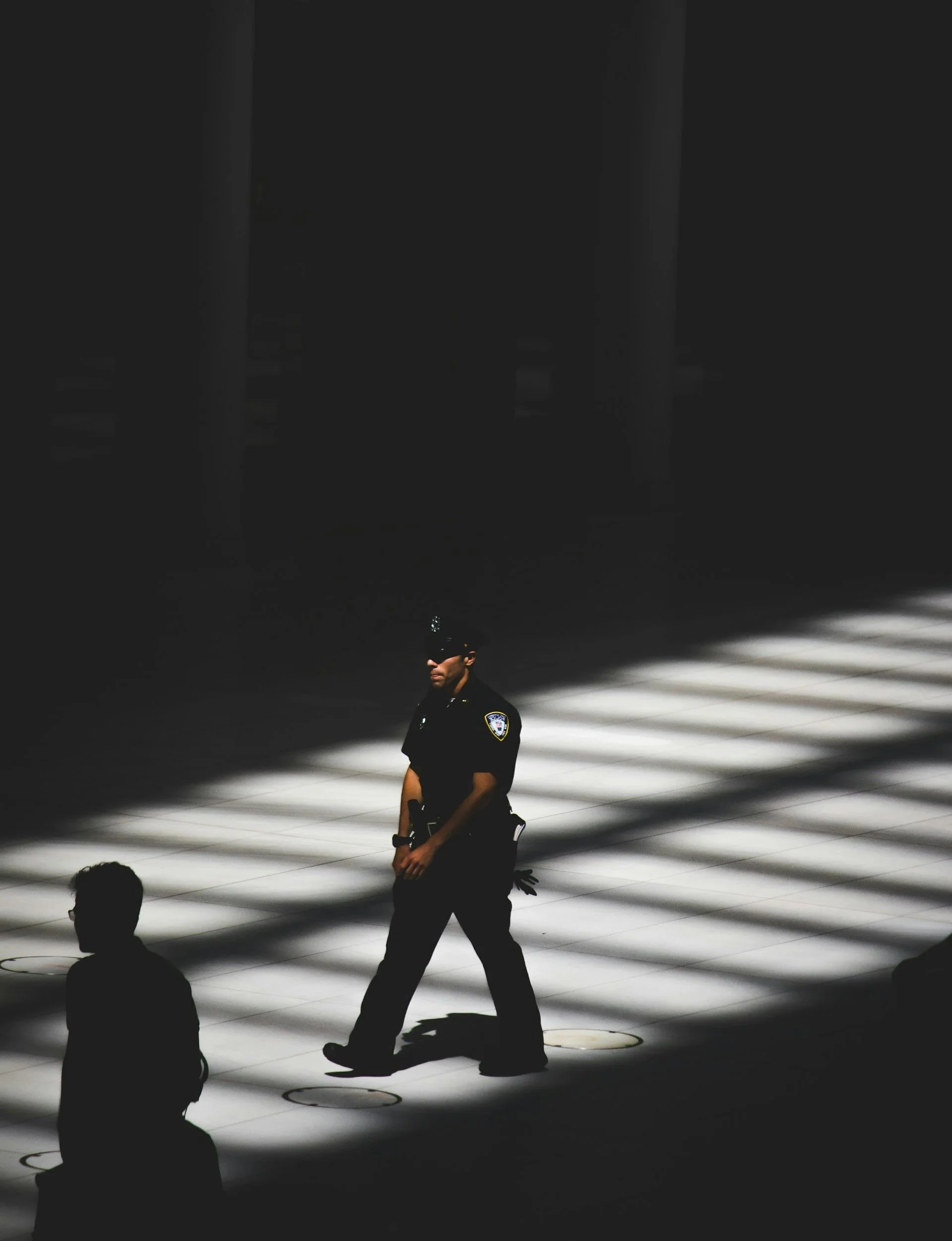 A police officer standing in a modern, minimalistic space with a man sitting on the floor and another person standing, both in silhouette. The scene features strong, directional lighting creating a stark contrast between light and shadow.