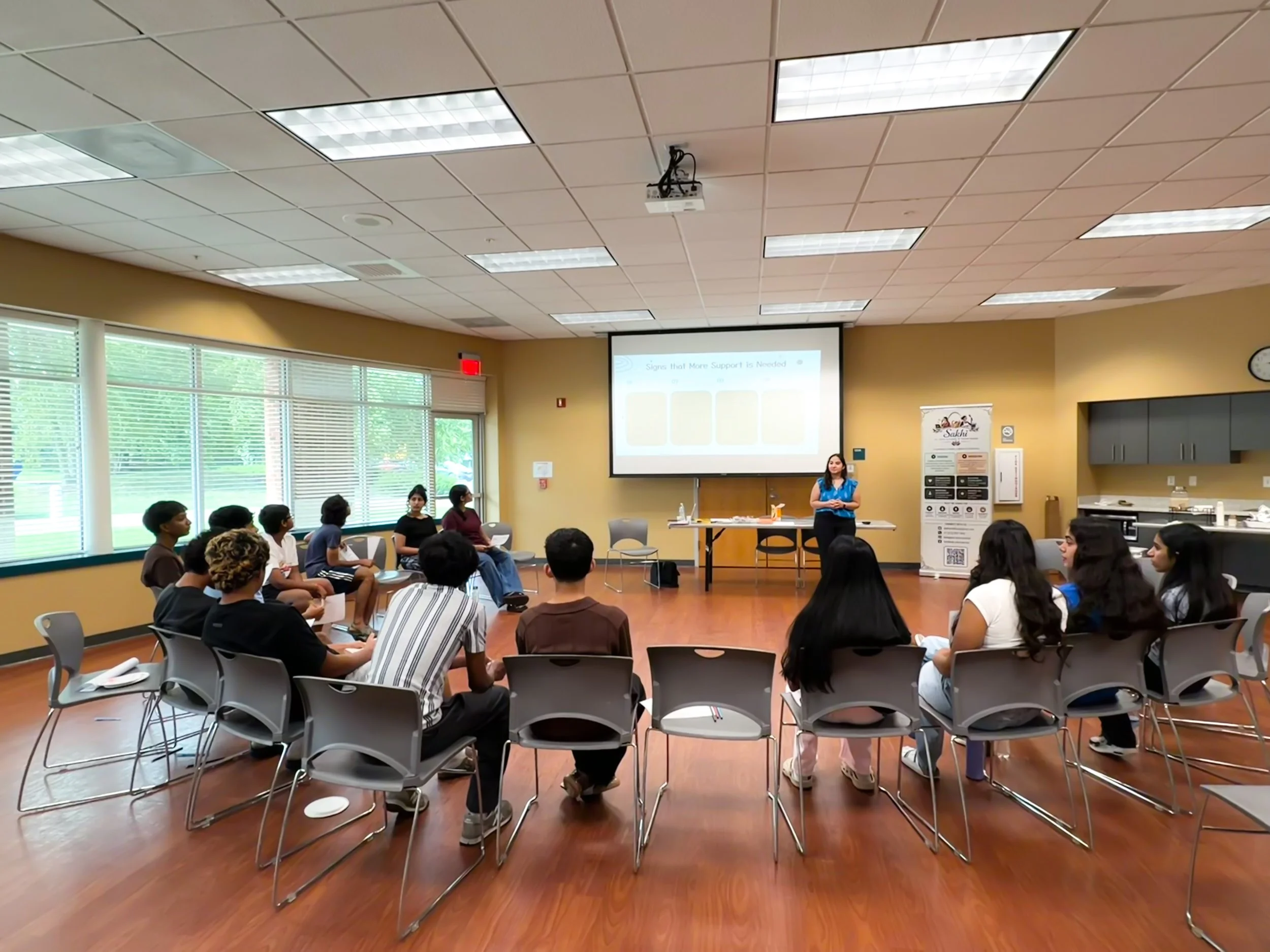 A classroom or seminar room with students seated in chairs facing a woman speaker. The woman is standing near a table at the front of the room, and there is a large screen displaying a presentation behind her. The room has large windows with blinds, beige walls, and a wooden floor.