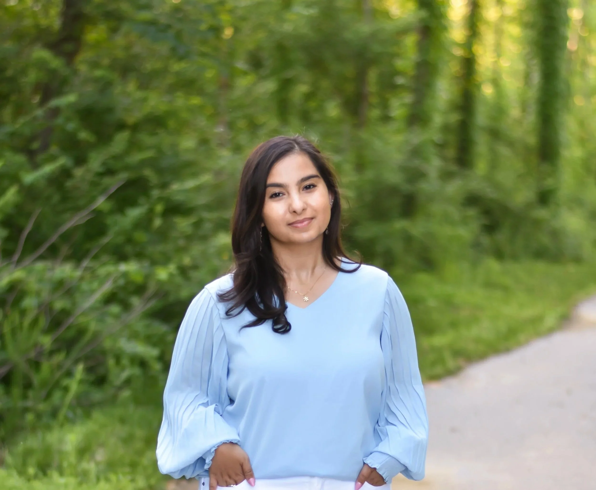 A young woman with long dark hair, wearing a light blue dress, standing outdoors in a green, leafy park with a pathway, smiling softly.