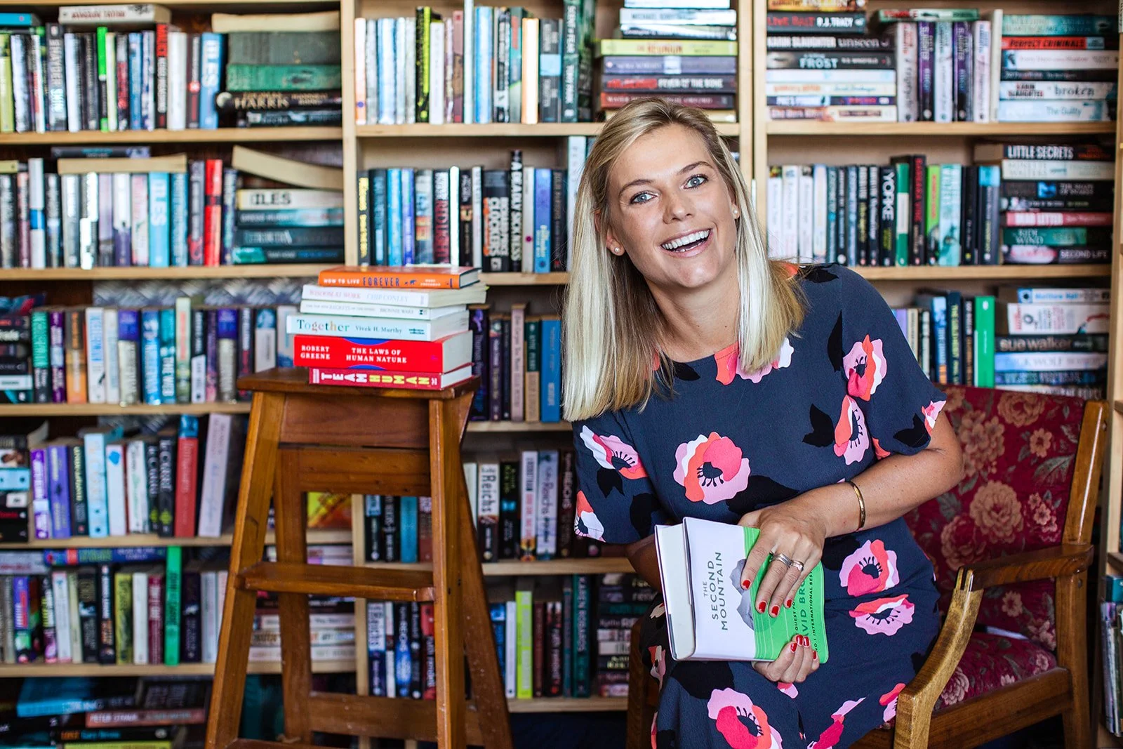 A woman with blonde hair smiling while sitting in a library filled with books, holding a book titled "The Second Mountain" by David Brooks.