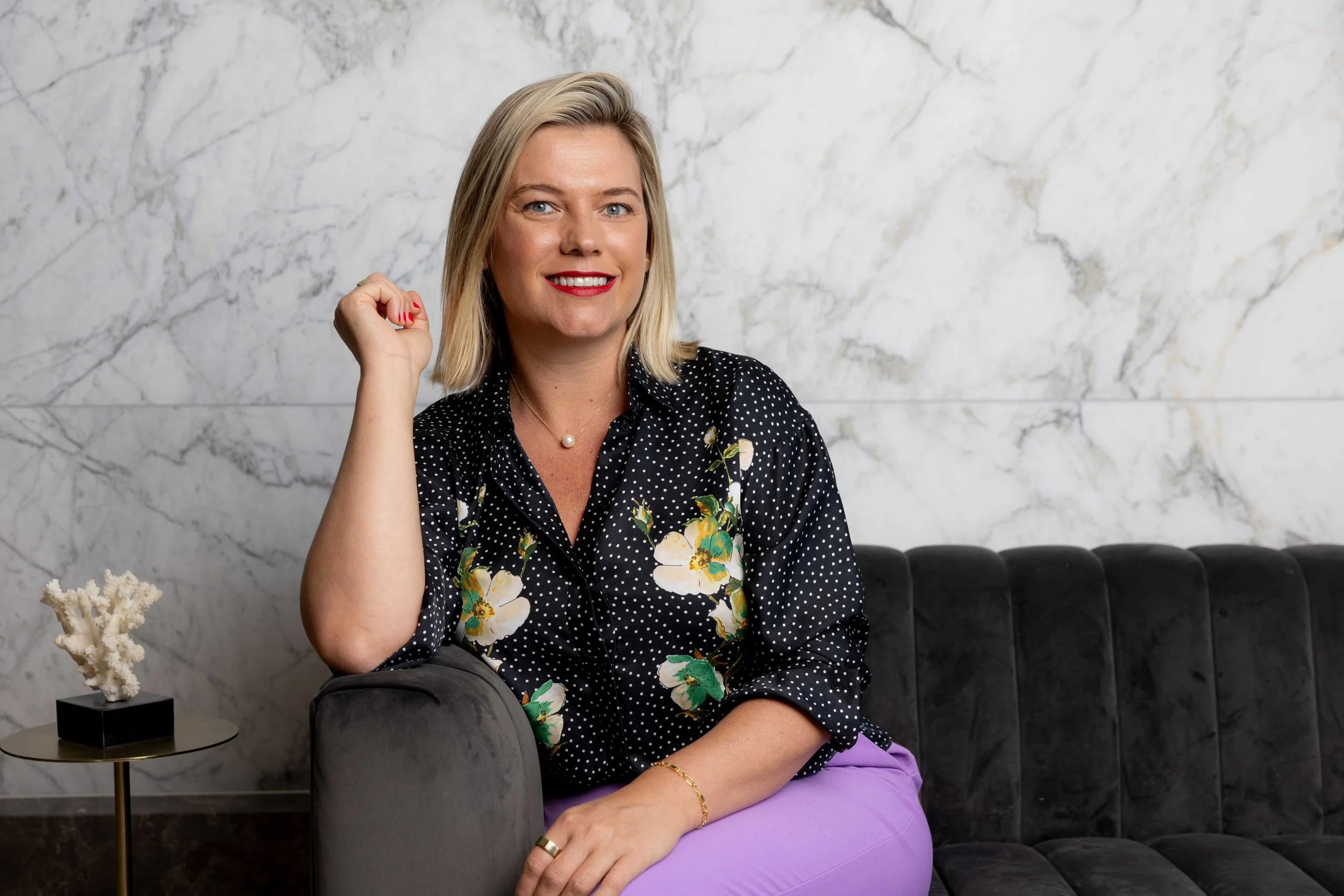 A woman with blonde hair, wearing a black polka dot shirt with floral print and purple pants, sitting on a black velvet sofa in front of a marble wall. She's smiling, with her right hand near her face, and has red lipstick and a pearl necklace.