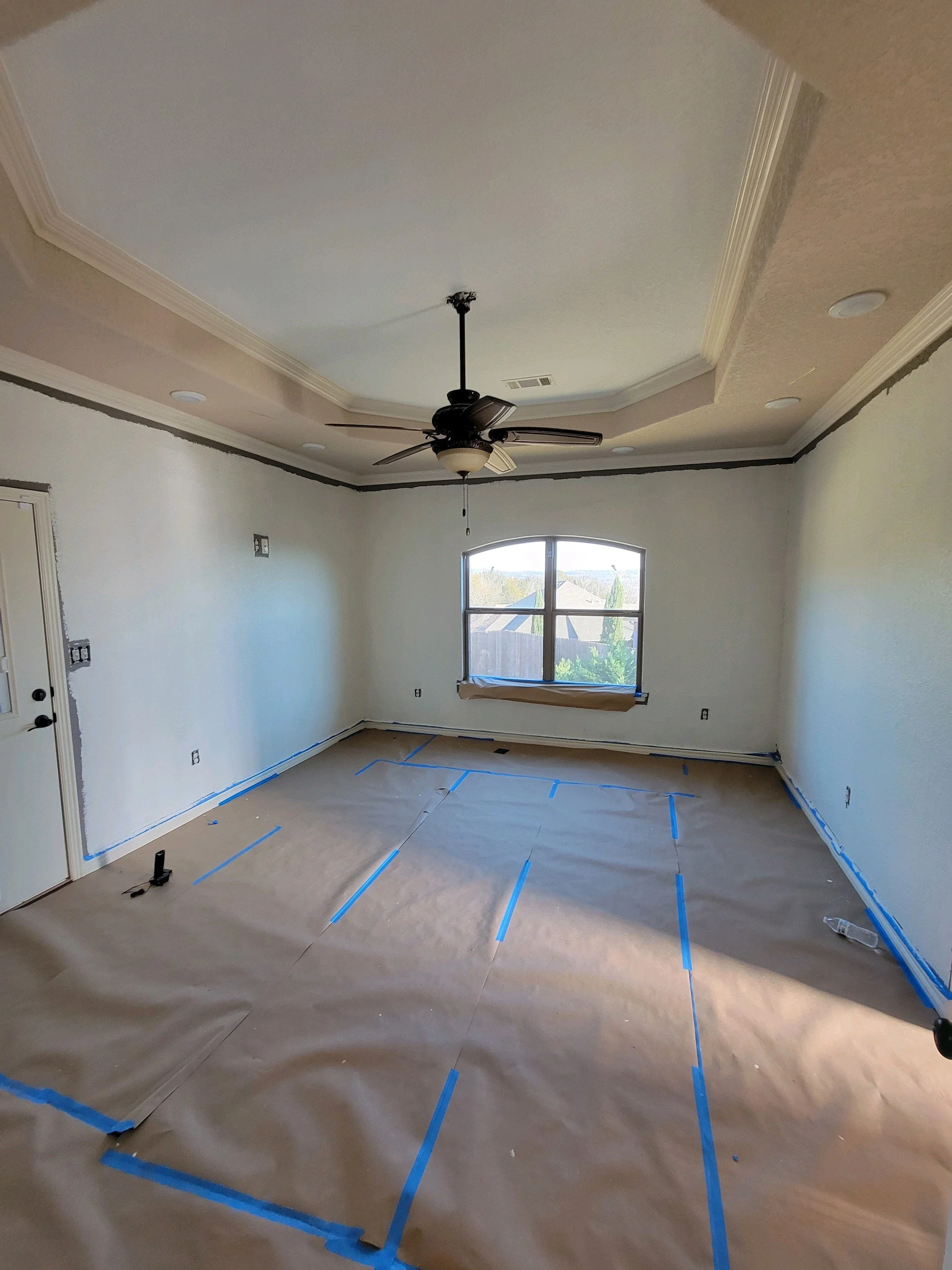 Room undergoing renovation with taped paper on floor and walls, ceiling fan installed, and window with view of rooftops and trees outside.