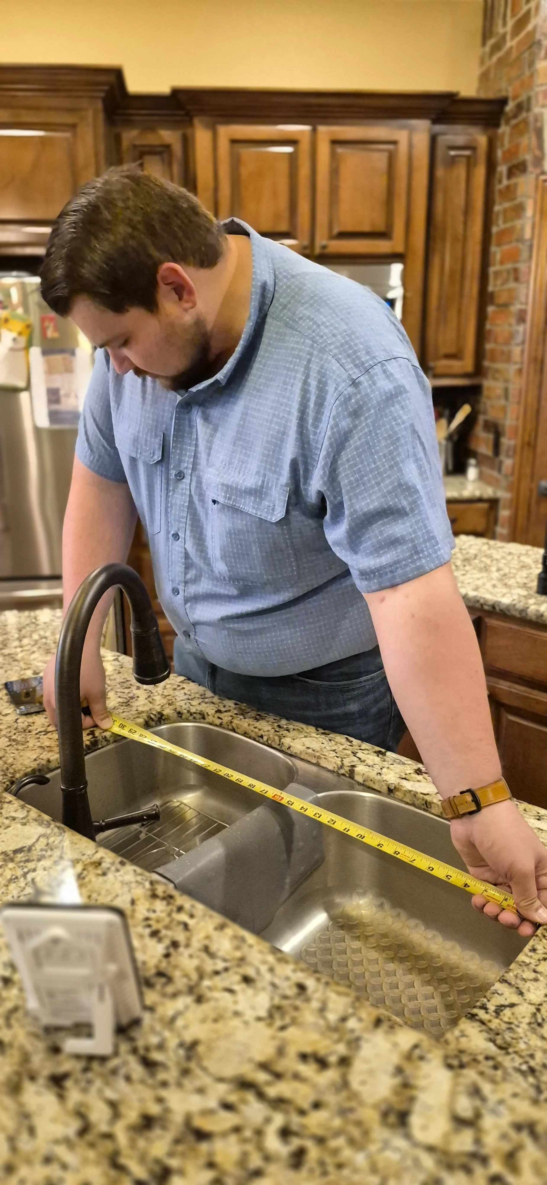 Abbott's Owner measuring a kitchen sink with a tape measure in a kitchen with wooden cabinets and granite countertops.