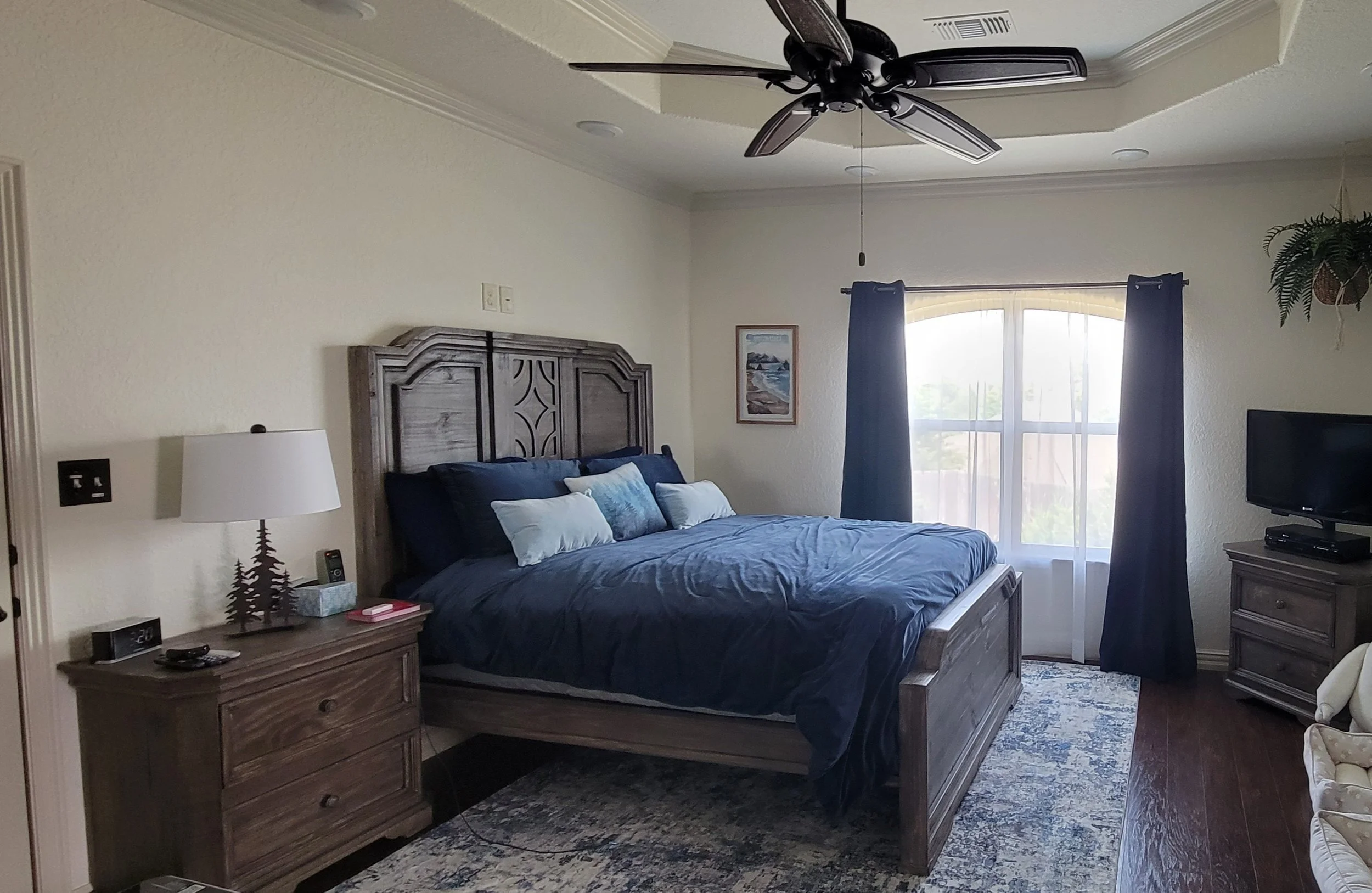 A bedroom with a wooden bed, blue bedding, dark curtains, a window with sheer curtains, a nightstand with a lamp and clock, a dresser with a TV, and a ceiling fan.