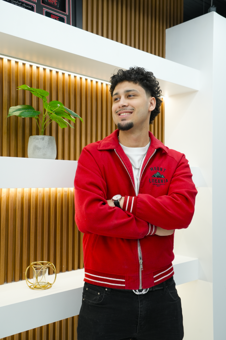 A young man with curly dark hair and a goatee, smiling and standing with his arms crossed in front of a white shelf. He is wearing a red jacket with a 'Mount Lucania' logo, a white T-shirt, black jeans, and a watch. The background features wooden paneling, a potted plant, and decorative objects.