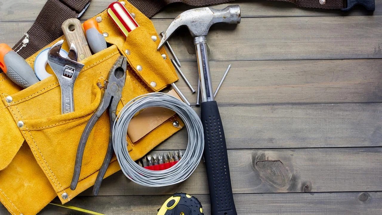 A yellow tool belt with several hand tools, including a wrench, pliers, and screwdrivers, on a wooden surface, with a coil of wire, a hammer, and other small tools nearby.
