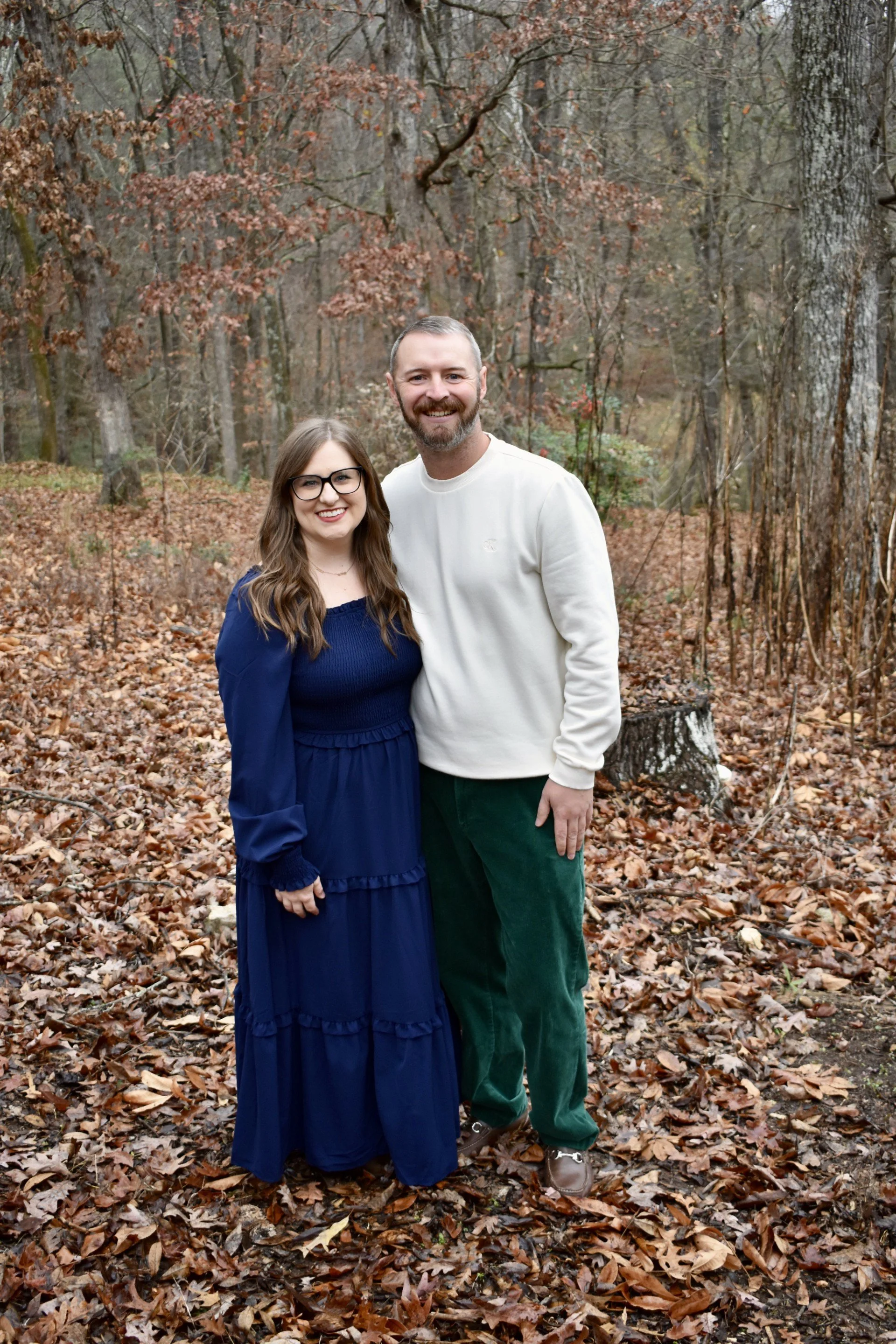 A smiling man and woman standing in a forest with fallen leaves and bare trees in autumn.
