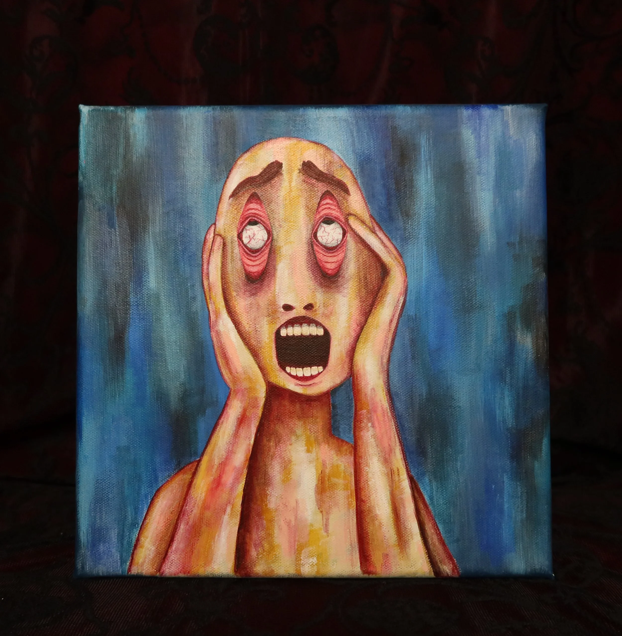 A painting of a screaming face with hands on its cheeks, surrounded by a blue background. The painting is on a small canvas resting on a dark surface.