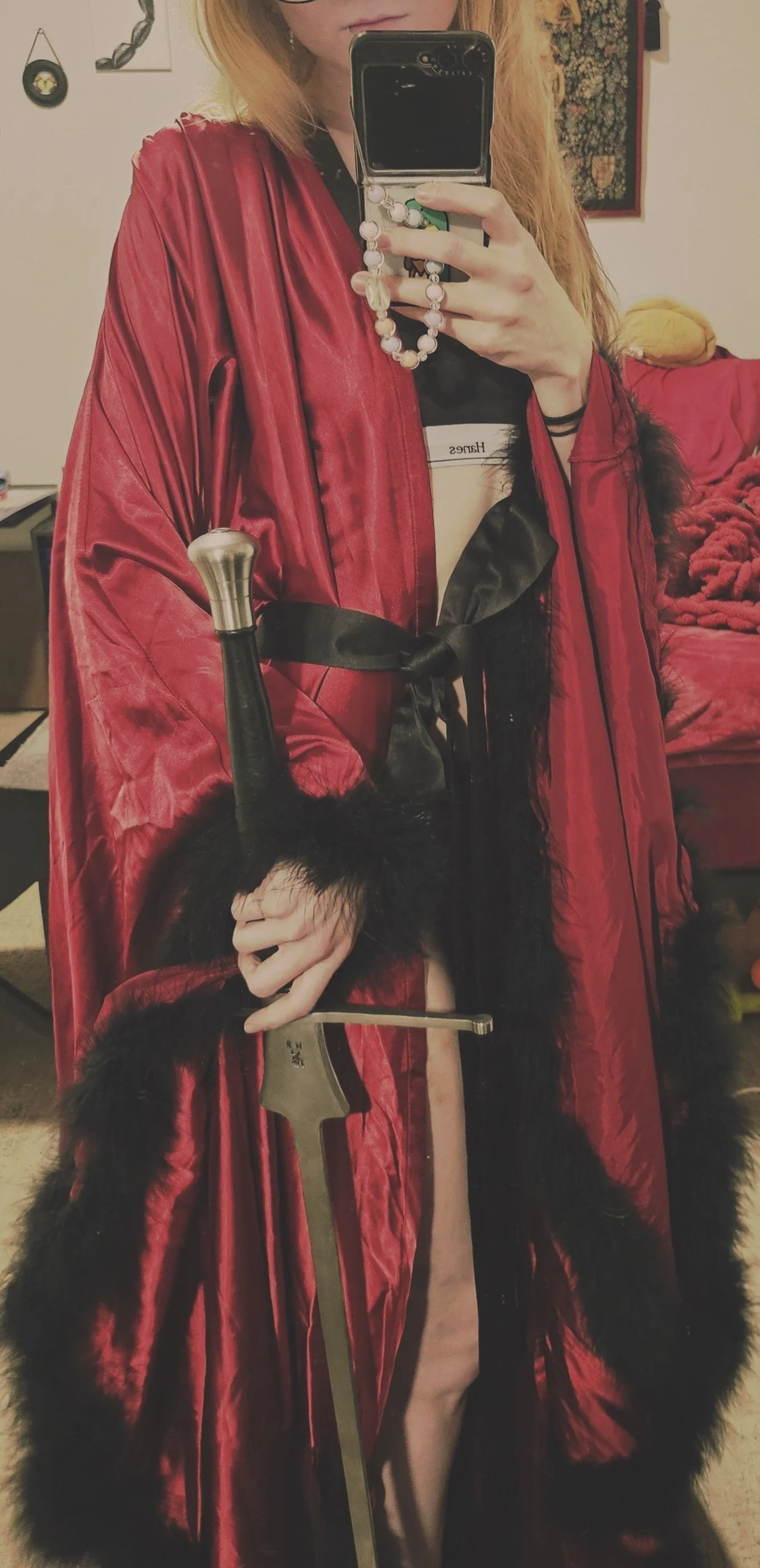 Person wearing a red cape with black fur trim, holding a sword, and standing in a room with a bed, wall art, and a mirror.