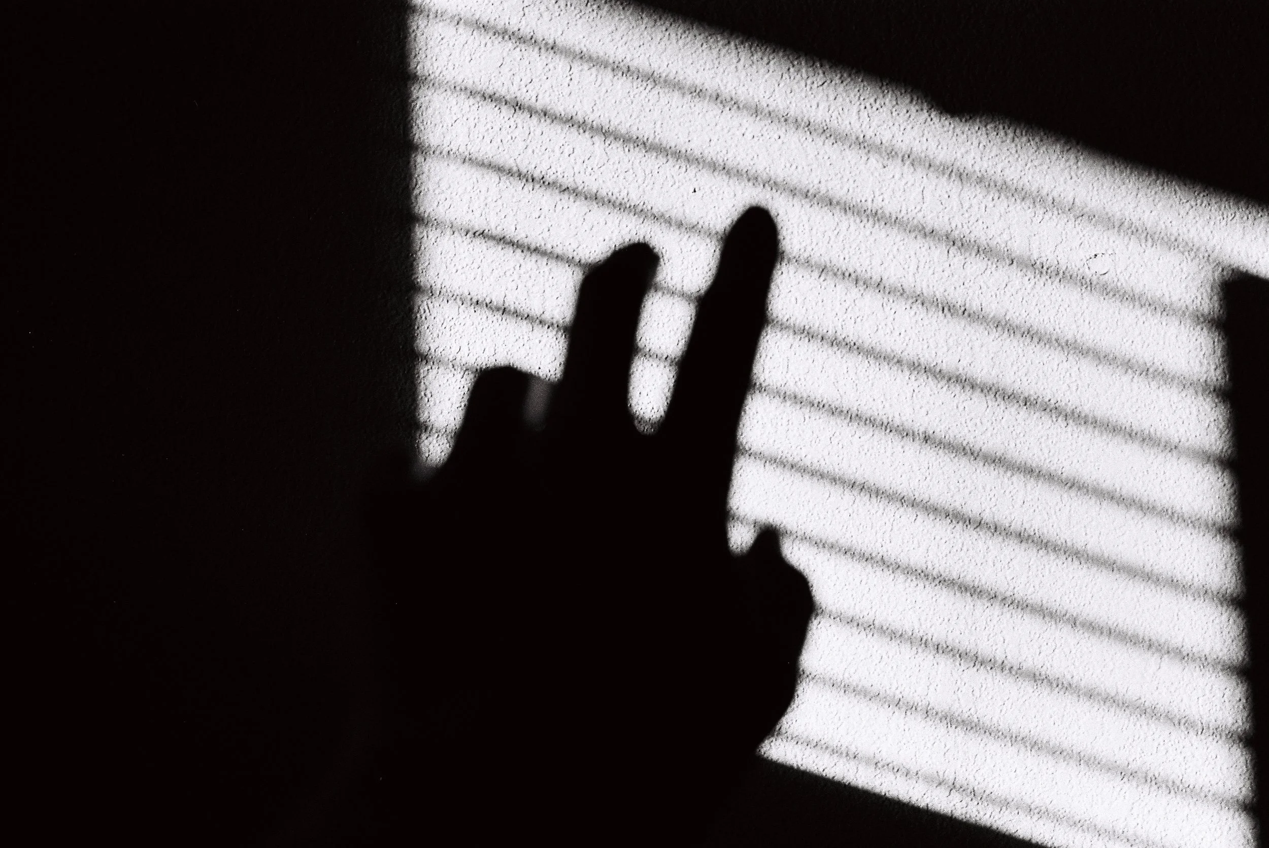 Silhouette of a hand reaching towards sunlight casting blinds on a wall.
