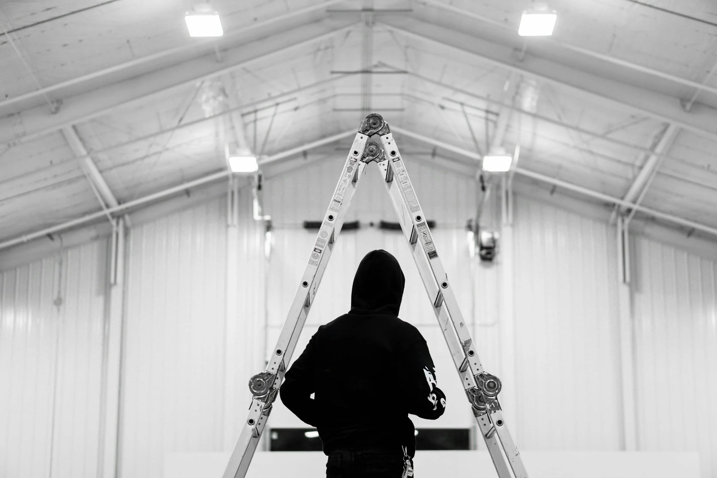 Person standing in an indoor space, facing away, with a ladder in front of them, metal beams and bright ceiling lights overhead.