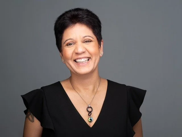 Smiling woman with short dark hair wearing a black top and a pendant necklace against a gray background.