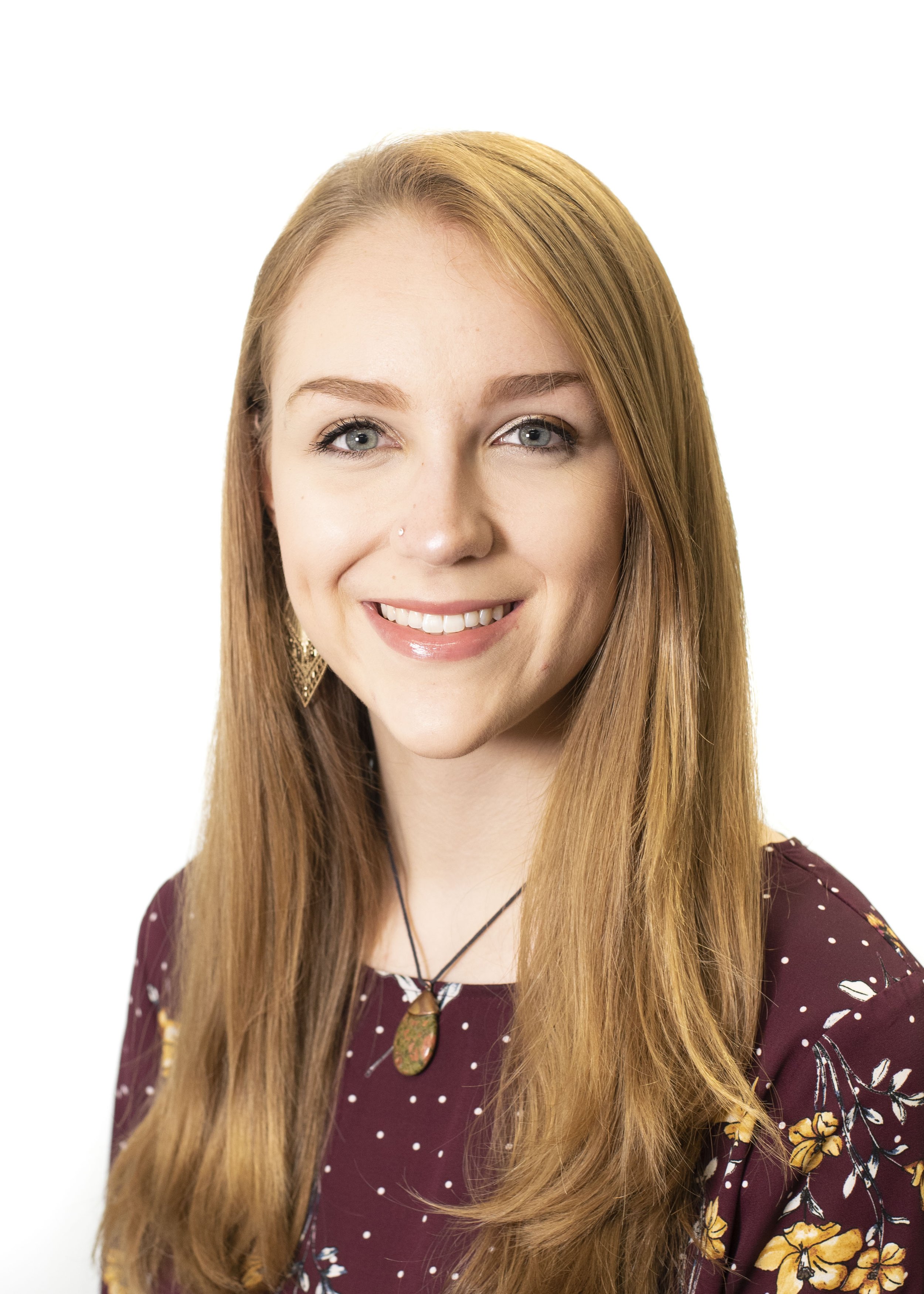 Close-up portrait of a young woman with long red hair, blue eyes, wearing a maroon dress with yellow floral patterns, and a pendant necklace, smiling against a white background.