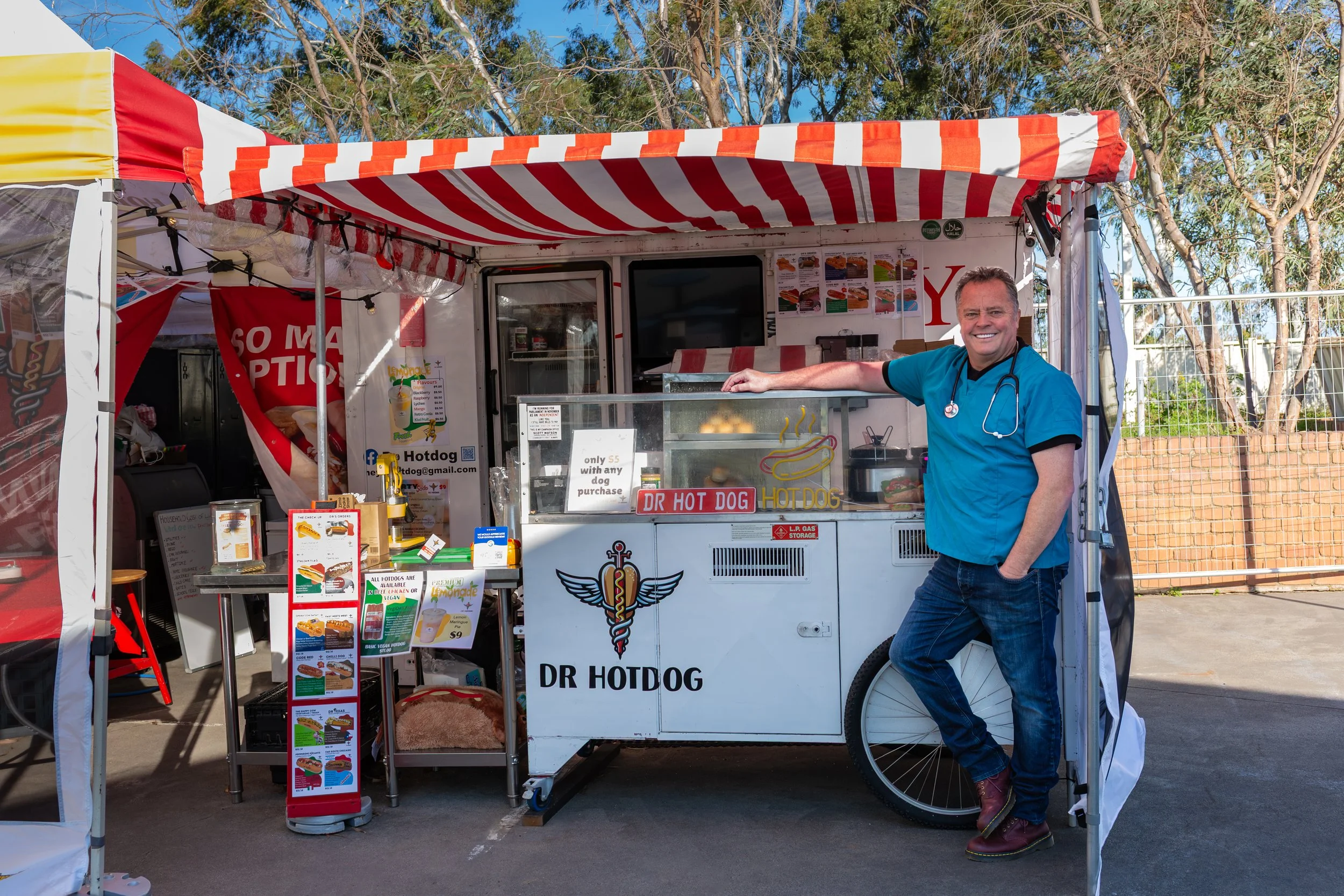A man in blue scrubs and jeans standing next to a white hot dog food cart with a red and white striped canopy, smiling at the camera. The cart displays menu items and signs, with a background of trees and a fence.