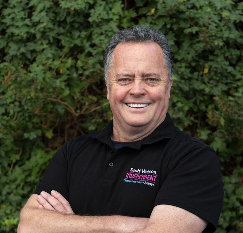 A smiling man standing outdoors in front of green bushes, wearing a black polo shirt with pink and white embroidered text, reading 'Scott Watson Independent'.