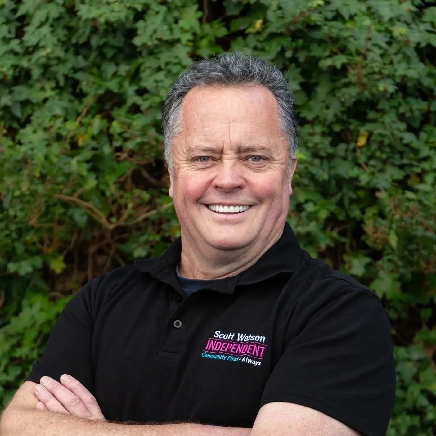 A smiling man, wearing a black polo shirt with embroidered text saying 'Scott Watson Independent', standing in front of green foliage.