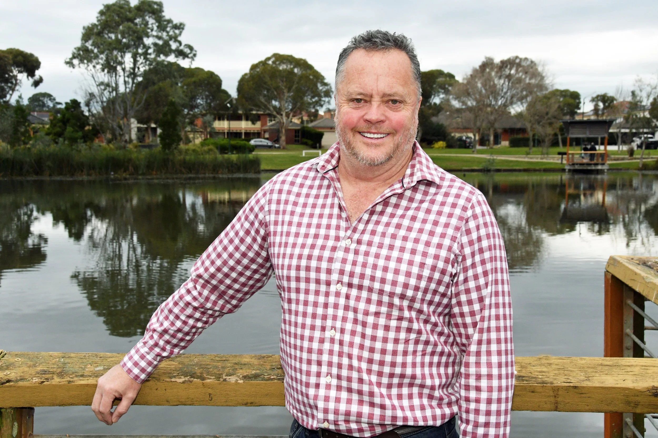 A smiling man in a red and white checkered shirt standing by a wooden railing near a pond, with houses and trees in the background under an overcast sky.