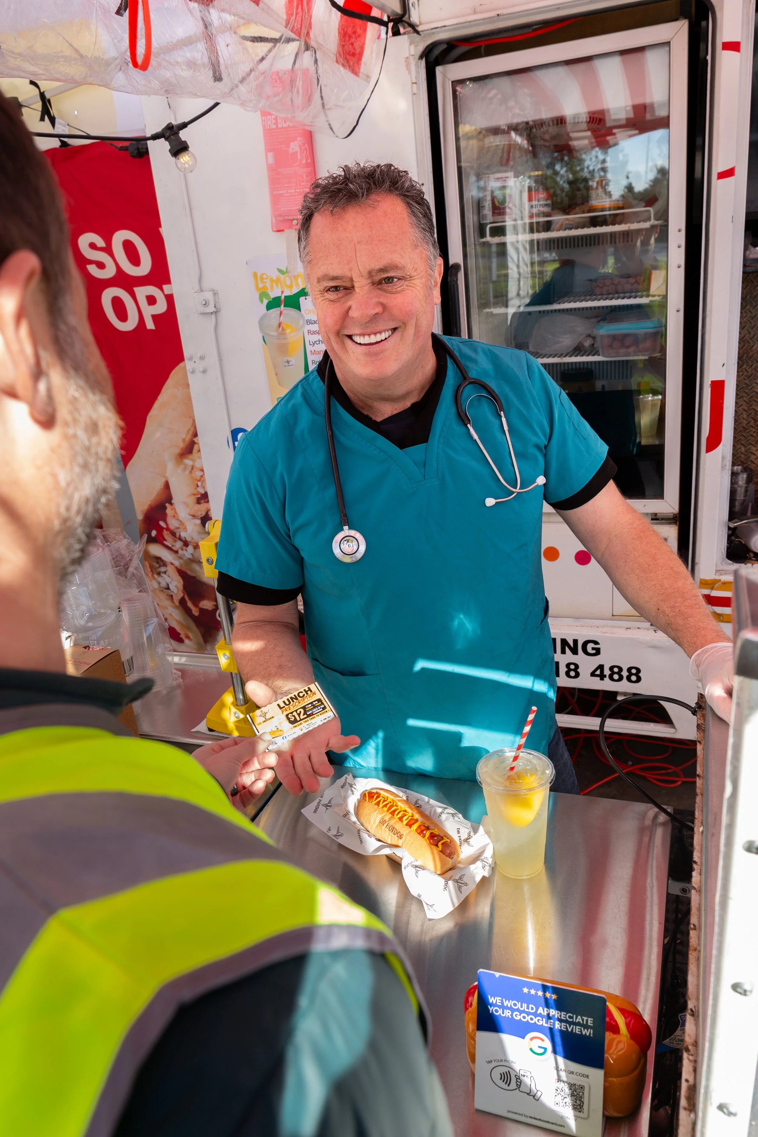 A man in medical scrubs with a stethoscope around his neck is smiling and selling hot dogs at a food stand. There is a hot dog with mustard and a beverage with a red and white striped straw on the counter.