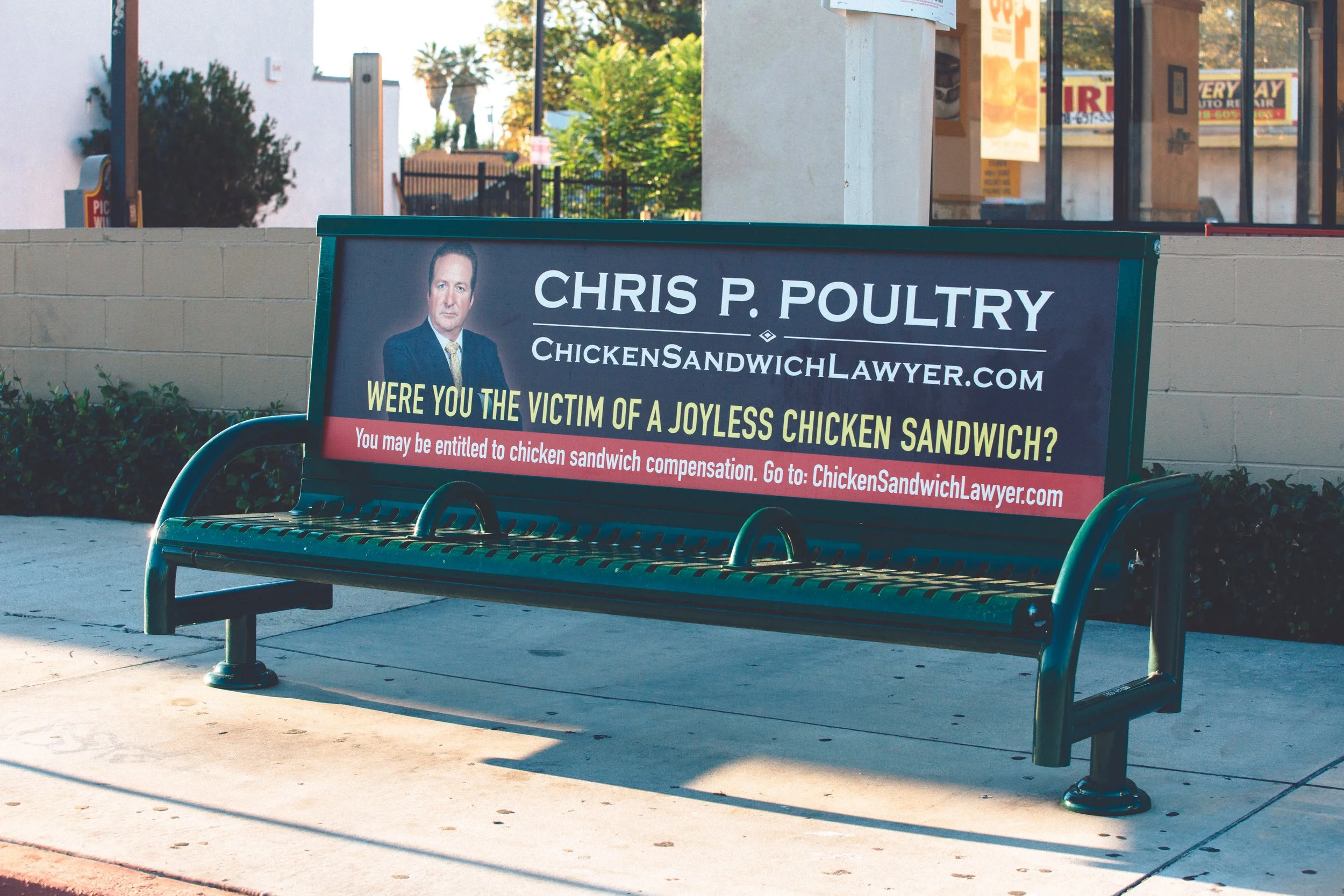A city street scene featuring a sidewalk and a green park bench with a political campaign sign. The sign promotes Chris P. Poultry for Chicken Sandwich Lawyer, asking if viewers were victims of a joyless chicken sandwich, and includes a website. In the background, there are buildings, trees, and a clear sky.