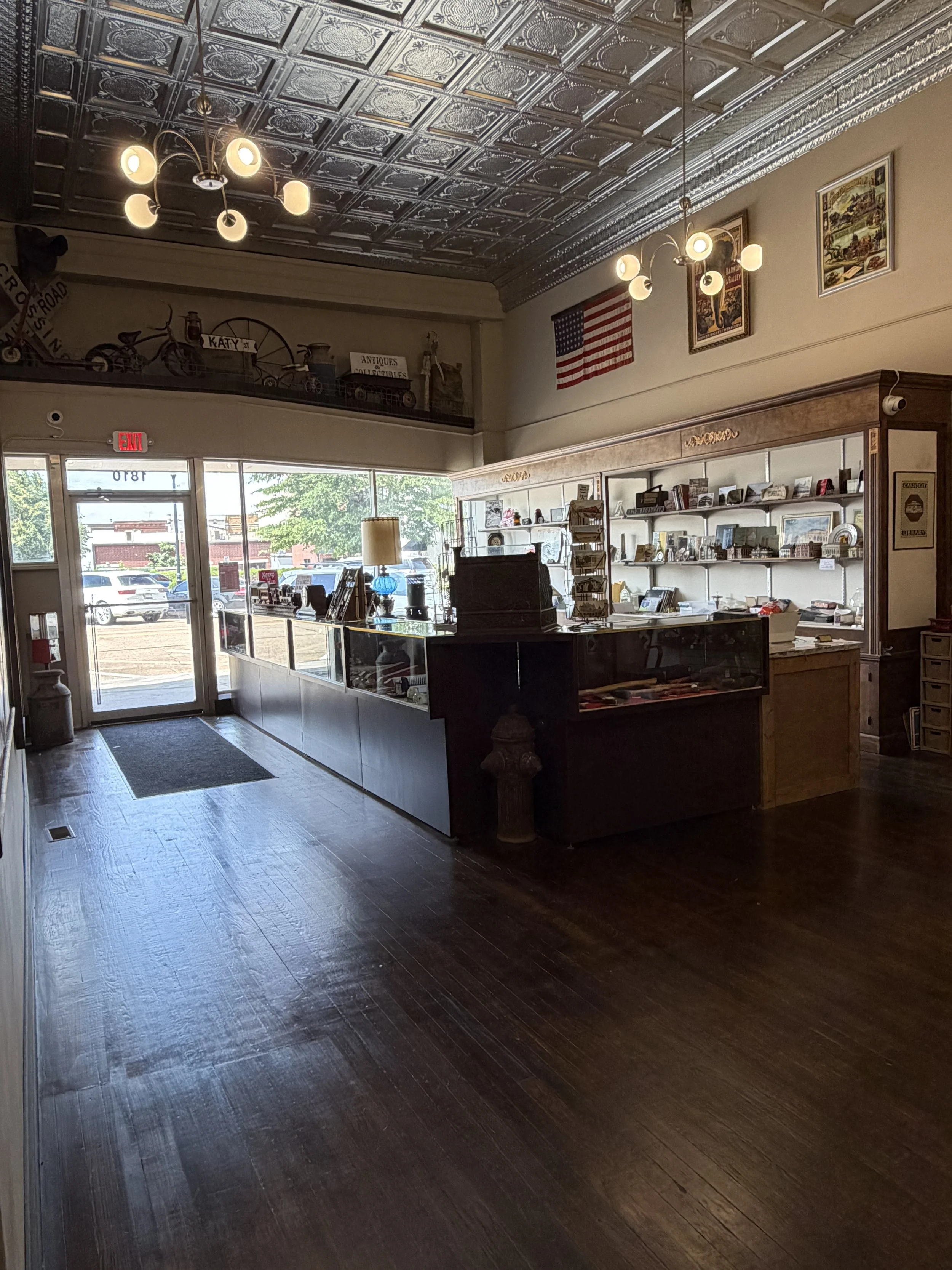 Inside of a vintage shop or antique store with a dark hardwood floor, glass display case, and white shelves filled with various collectibles. The front entrance with glass doors and windows allows natural light to illuminate the space. Metal signs and decorative items, including an American flag, are displayed on the wall and shelves.