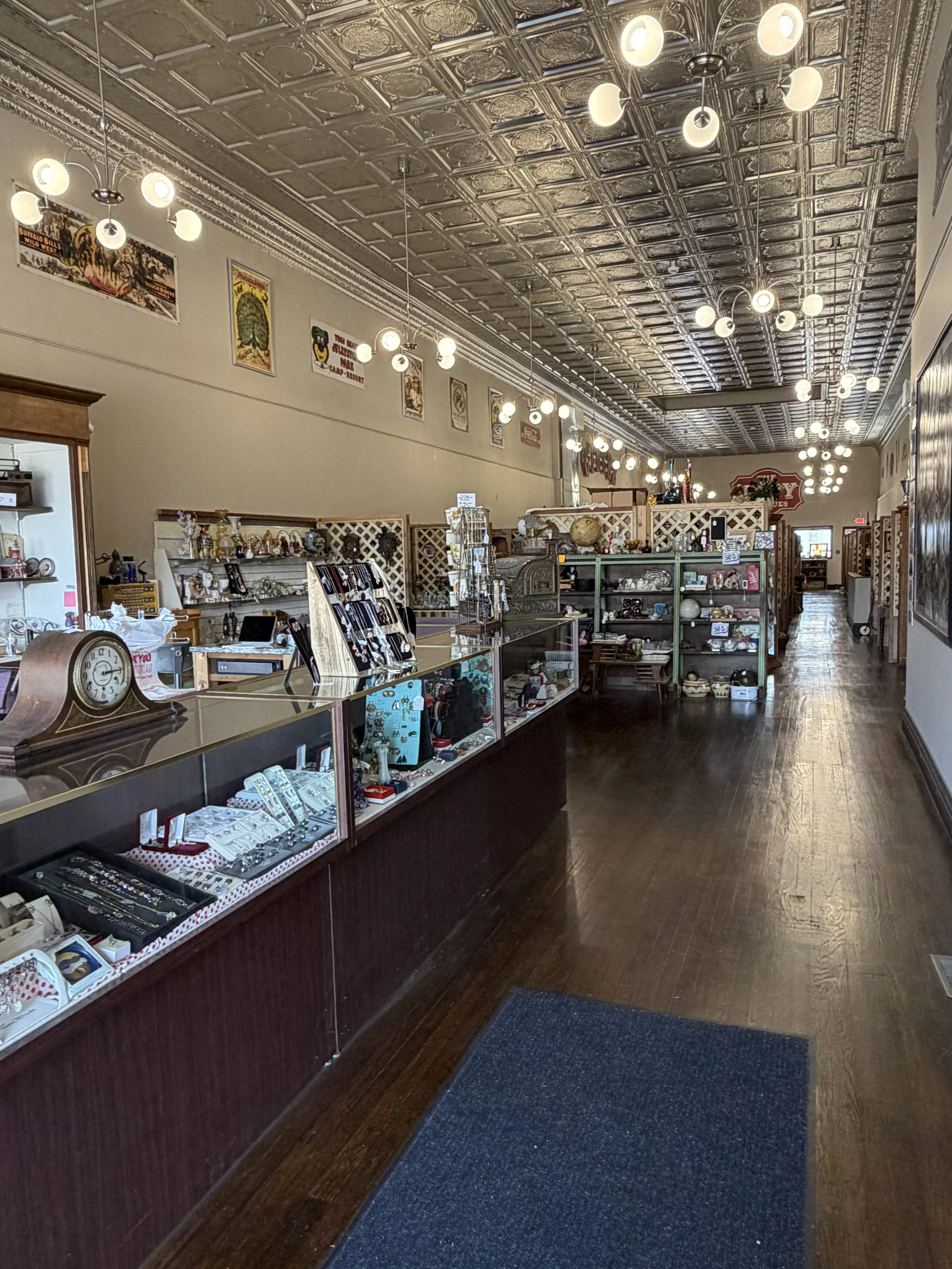 Interior of a vintage store with display cases filled with jewelry and collectibles, decorative posters on the wall, shiny tin ceiling, wooden flooring, and multiple hanging light fixtures.