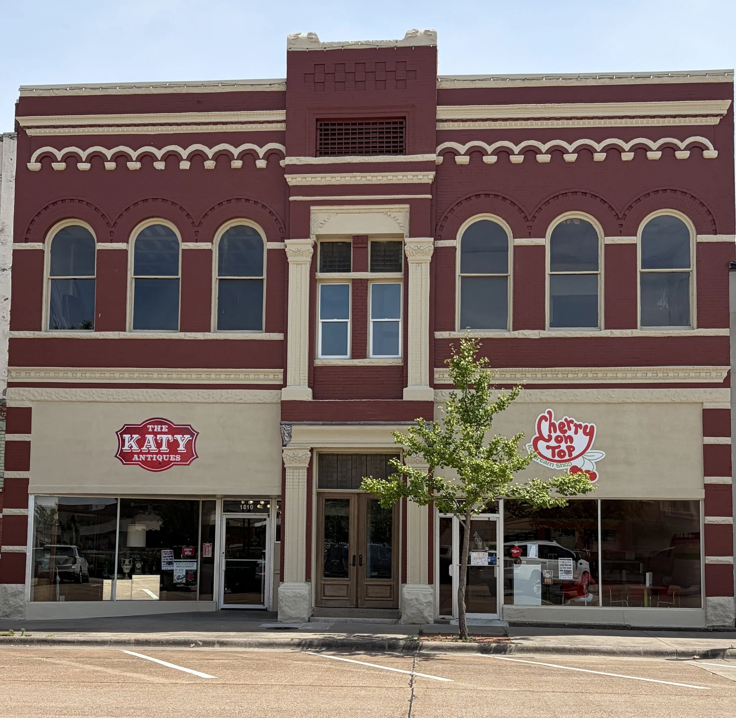 Red brick building with decorative white trim, arched windows, and signs for 'The Katy Antiques' and 'Cherry on Top Cream Shop', two small businesses on the ground floor.