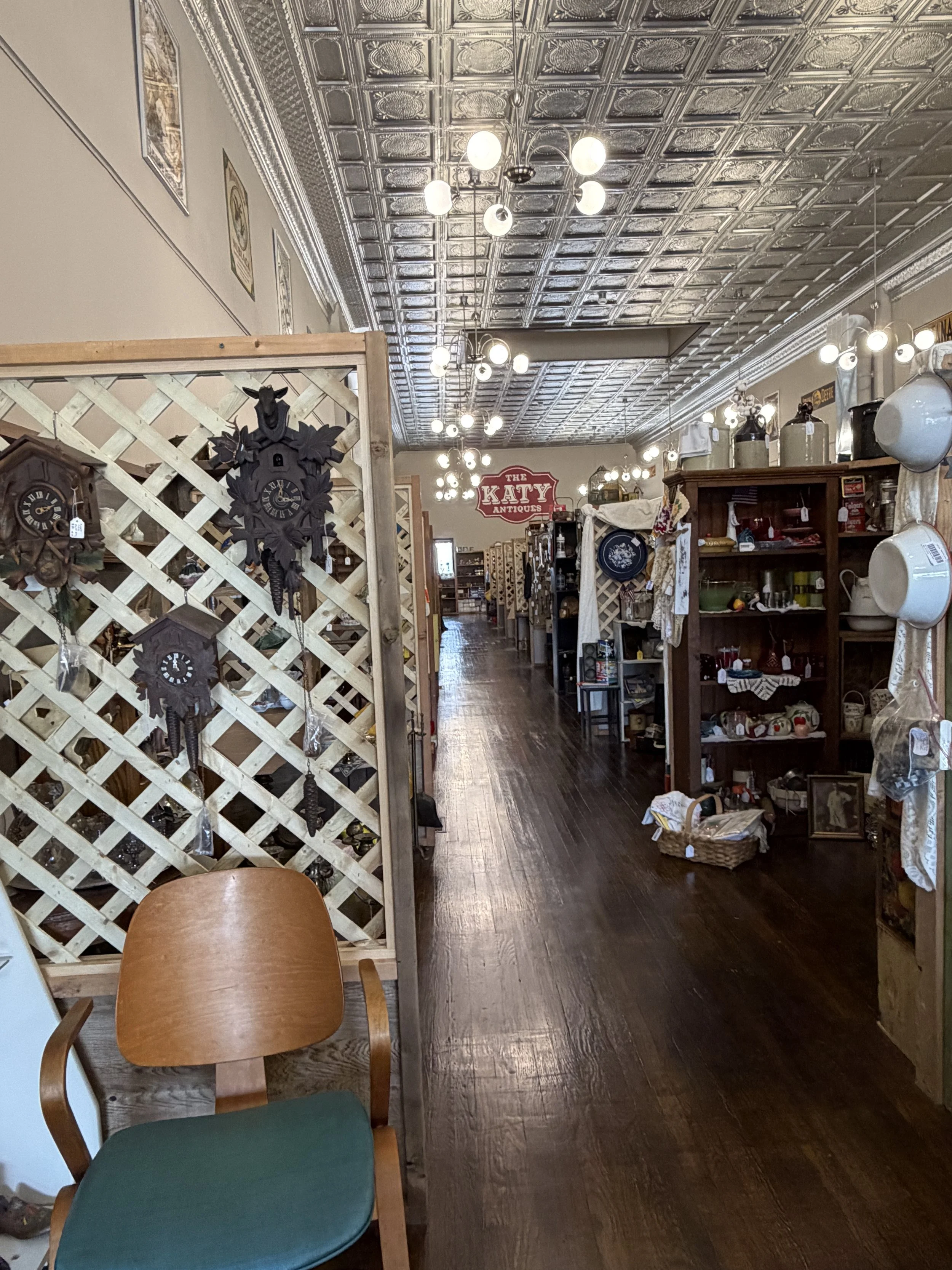 Interior of a vintage antique store with wooden shelves filled with various items, decorative lighting fixtures on a metallic tin ceiling, and a wooden lattice partition with antique clocks and wall decor. A sign reads "The Katy Antiques." There is a wooden chair with a green cushion in the foreground.