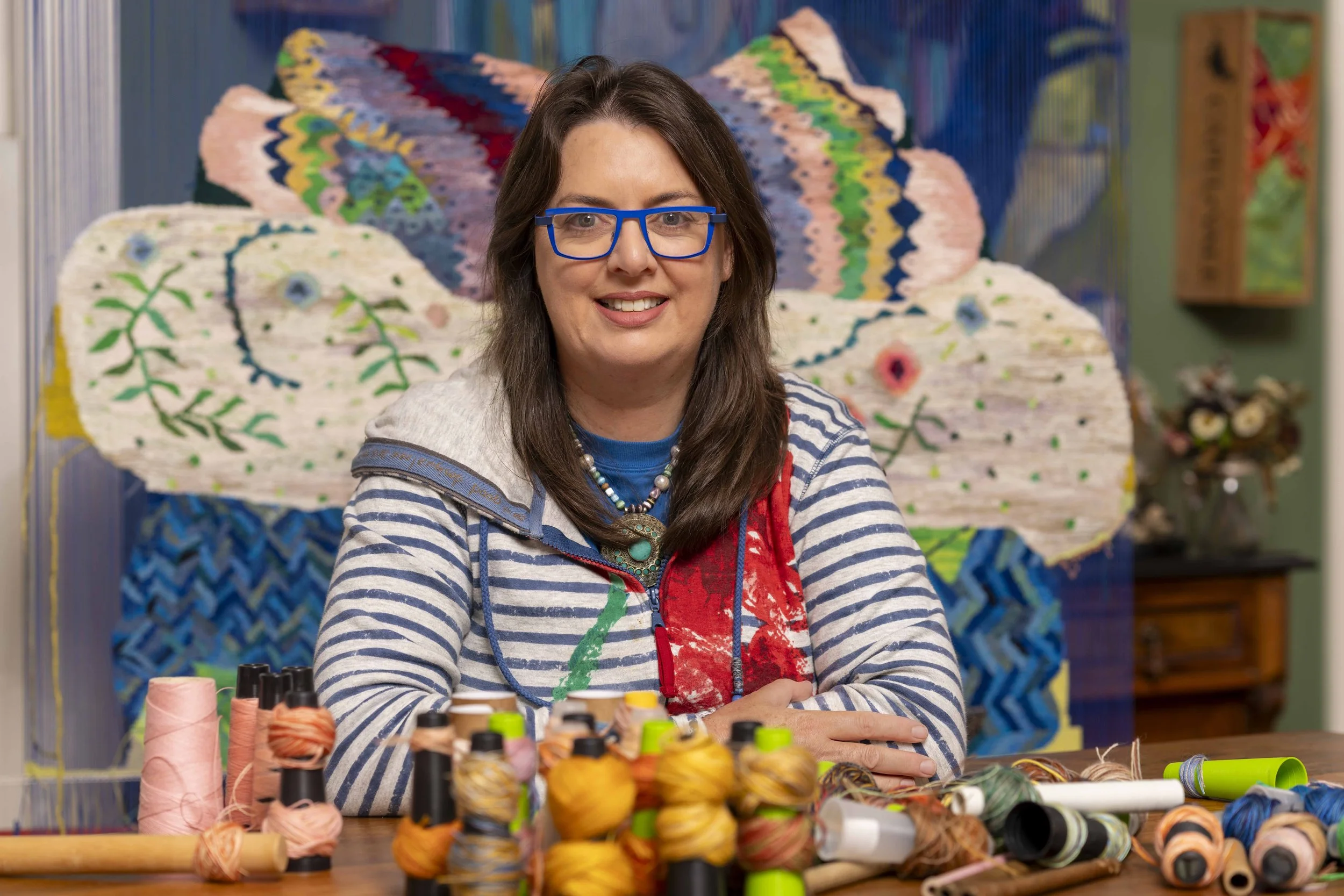 Dimity Kidston, wearing blue glasses, and a striped hoodie sitting at a table with colorful yarn and thread spools in the foreground, smiling at the camera, with a vibrant and colorful tapestry weaving hanging behind her.