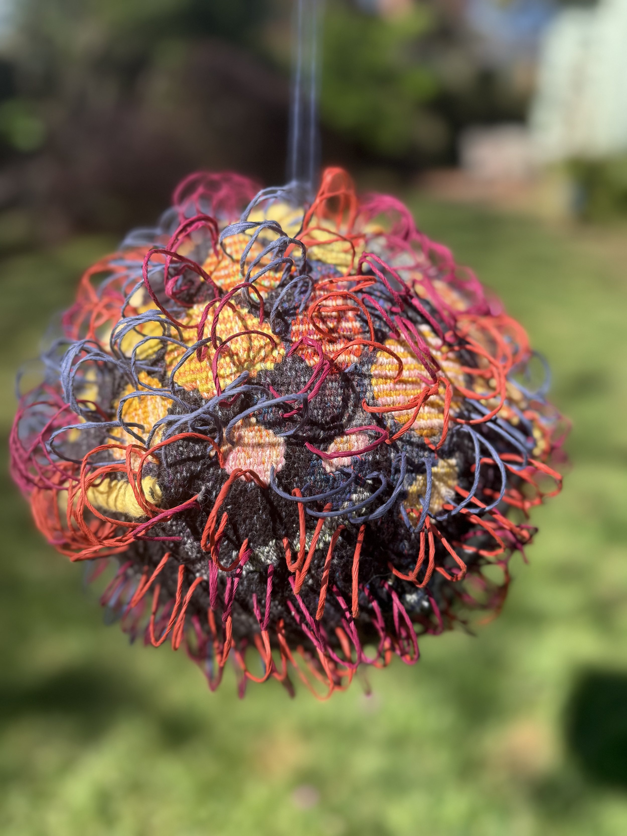 Close-up of a colorful ball of 3 dimensional woven tapestry with a blurred outdoor background.