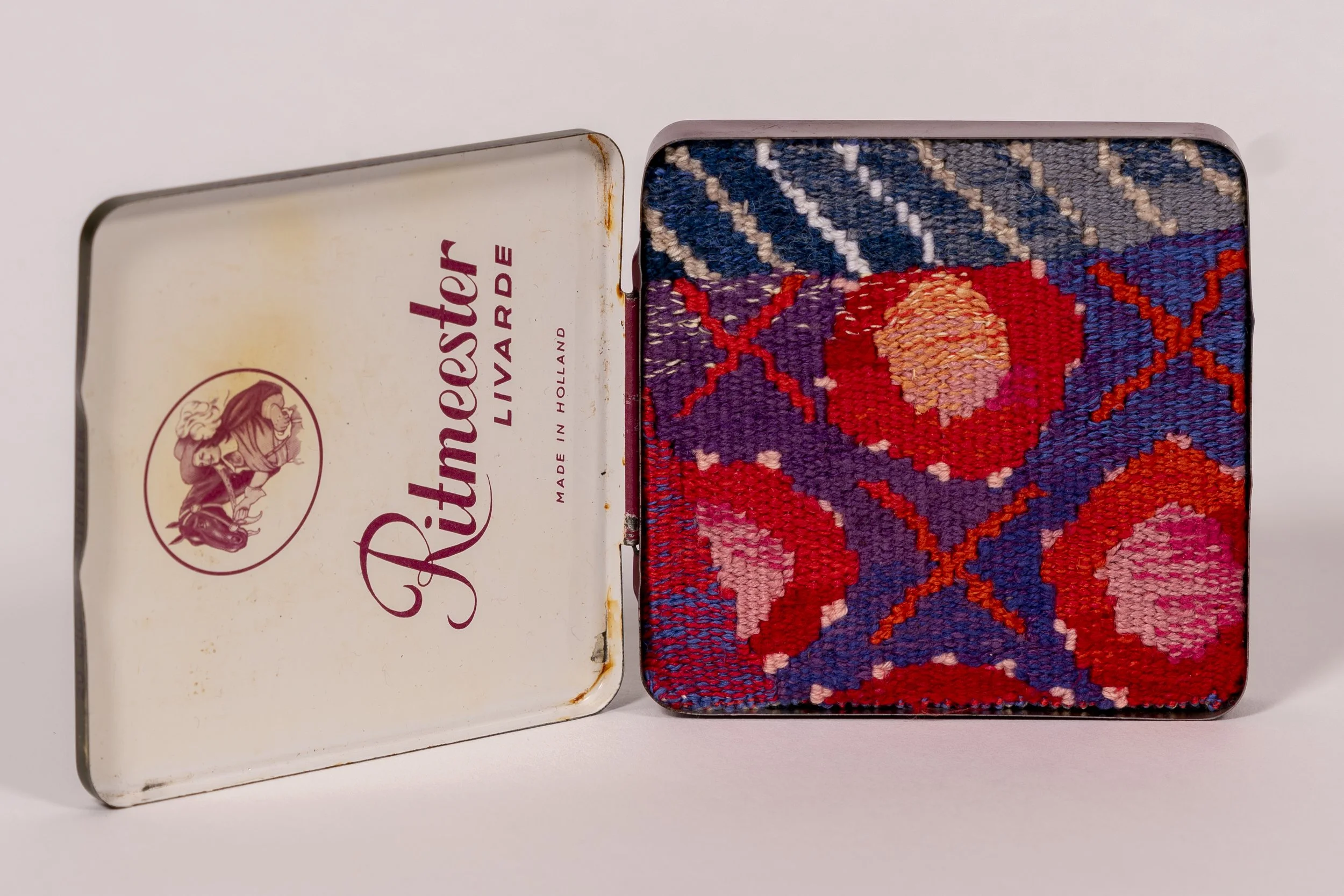 An open vintage metal tin box with a colorful embroidered design on the right side and a white lid with red lettering and a lion logo on the left side.