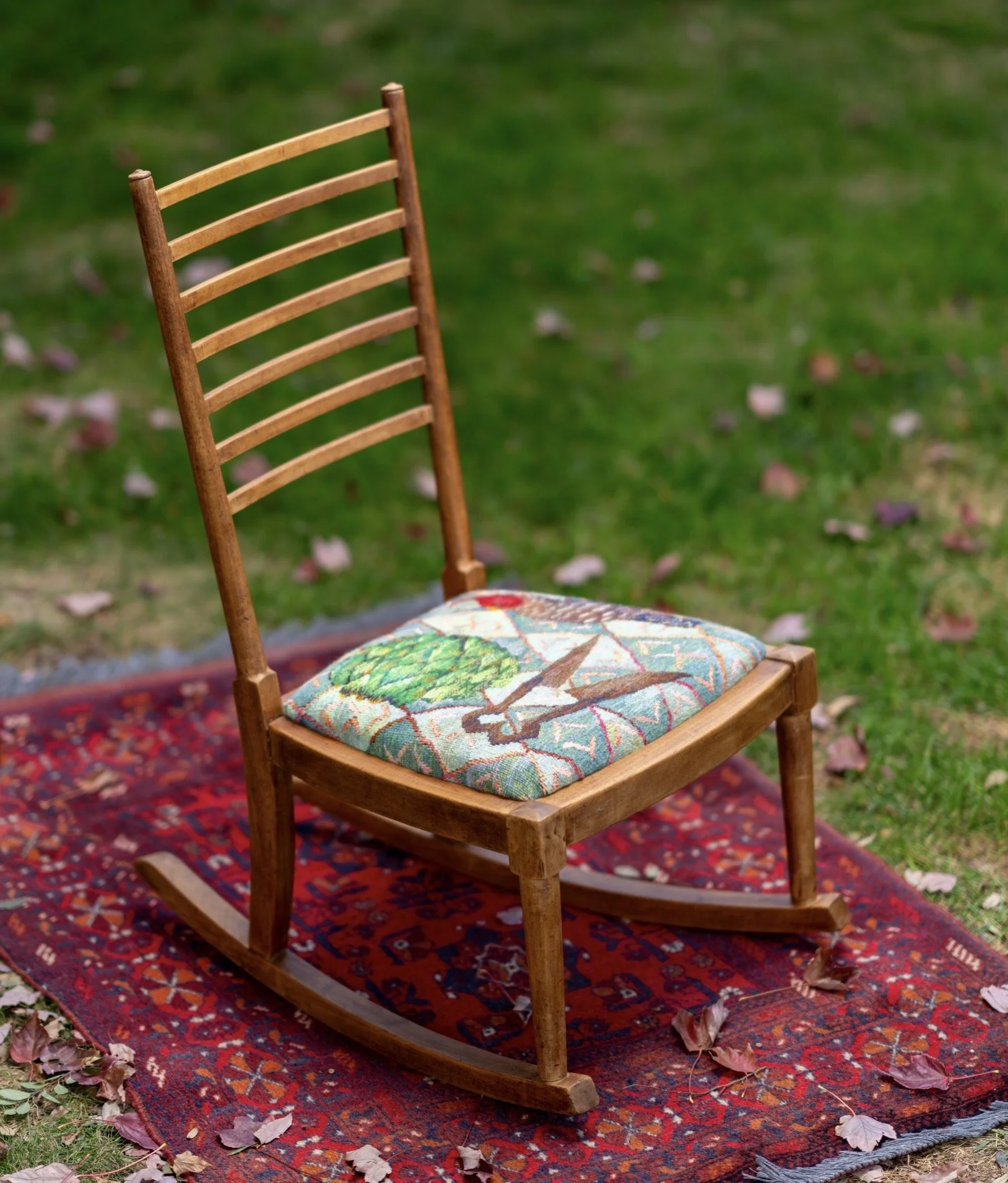 A wooden rocking chair with a floral cushion placed on a colorful rug outside, on grassy ground with fallen leaves.