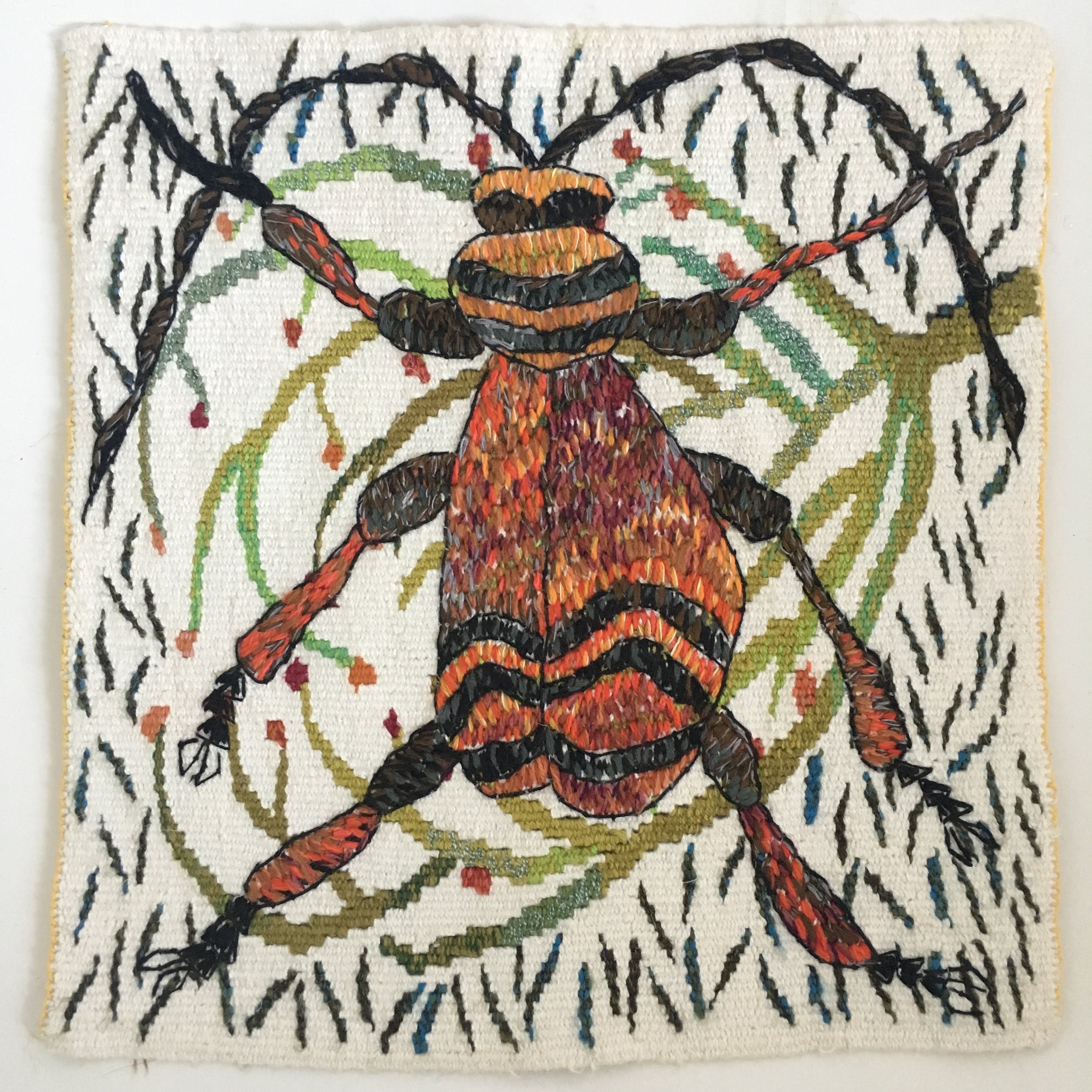 Tapestry weaving and embroidery of a beatle with orange, black, and yellow stripes, black wings, and legs, surrounded by green, red, and black patterns on a white fabric background.