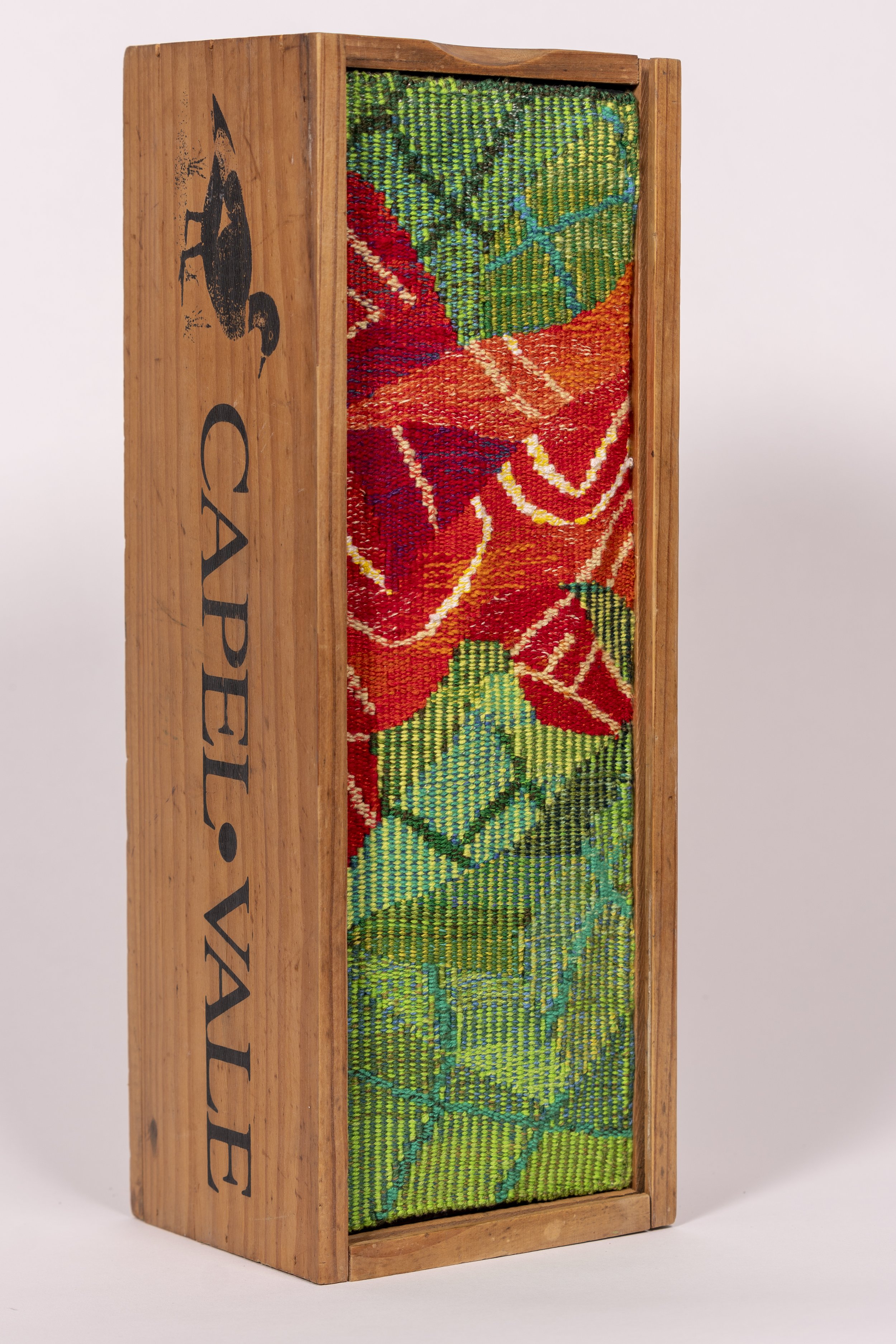 A wooden wine box with a woven tapestry on its front panel, featuring red, green, blue, and orange geometric patterns, and the word Capelvale printed on the side.