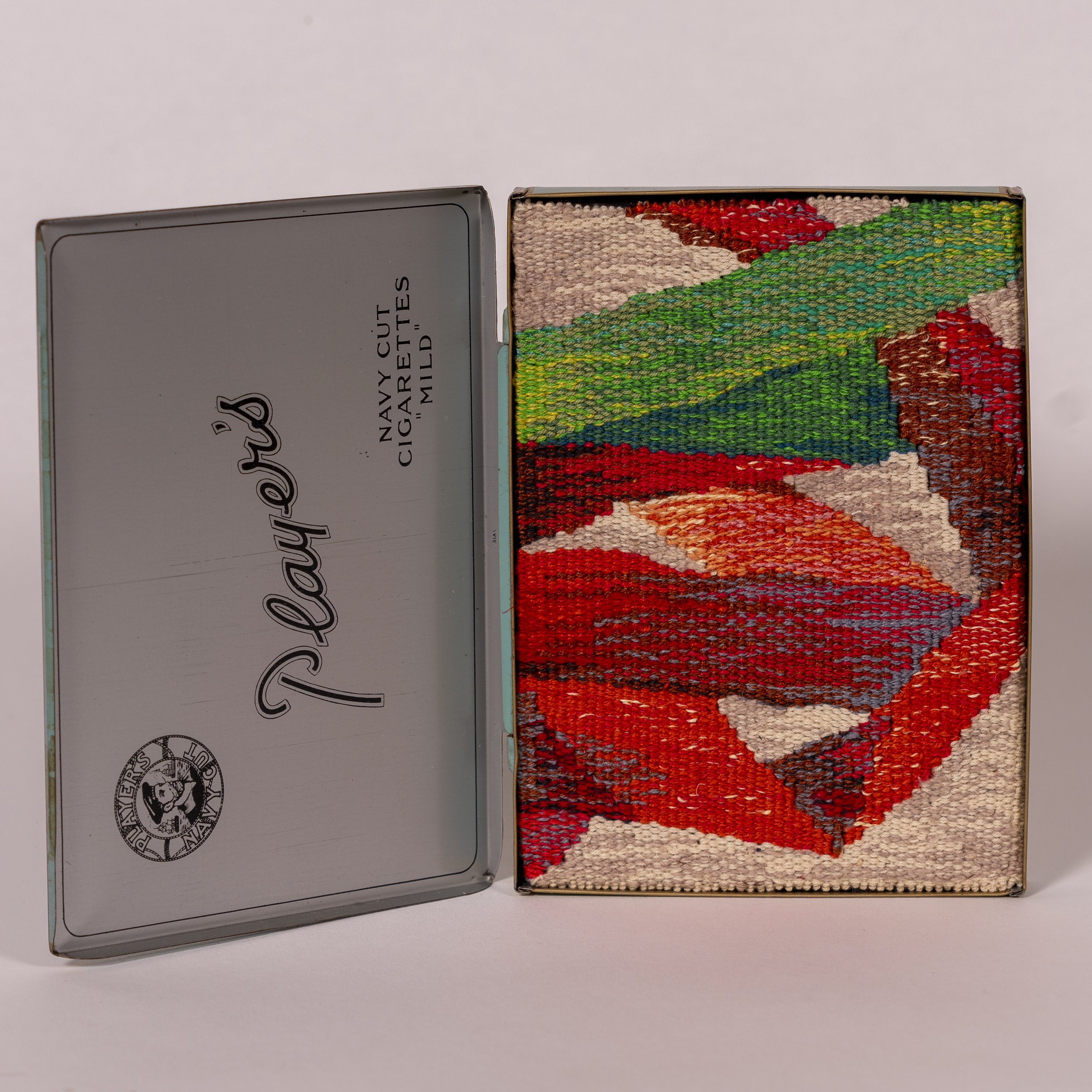 Open metal box with colorful embroidery of a mountain landscape scene inside, and a separate metal lid with partially visible text and a logo.