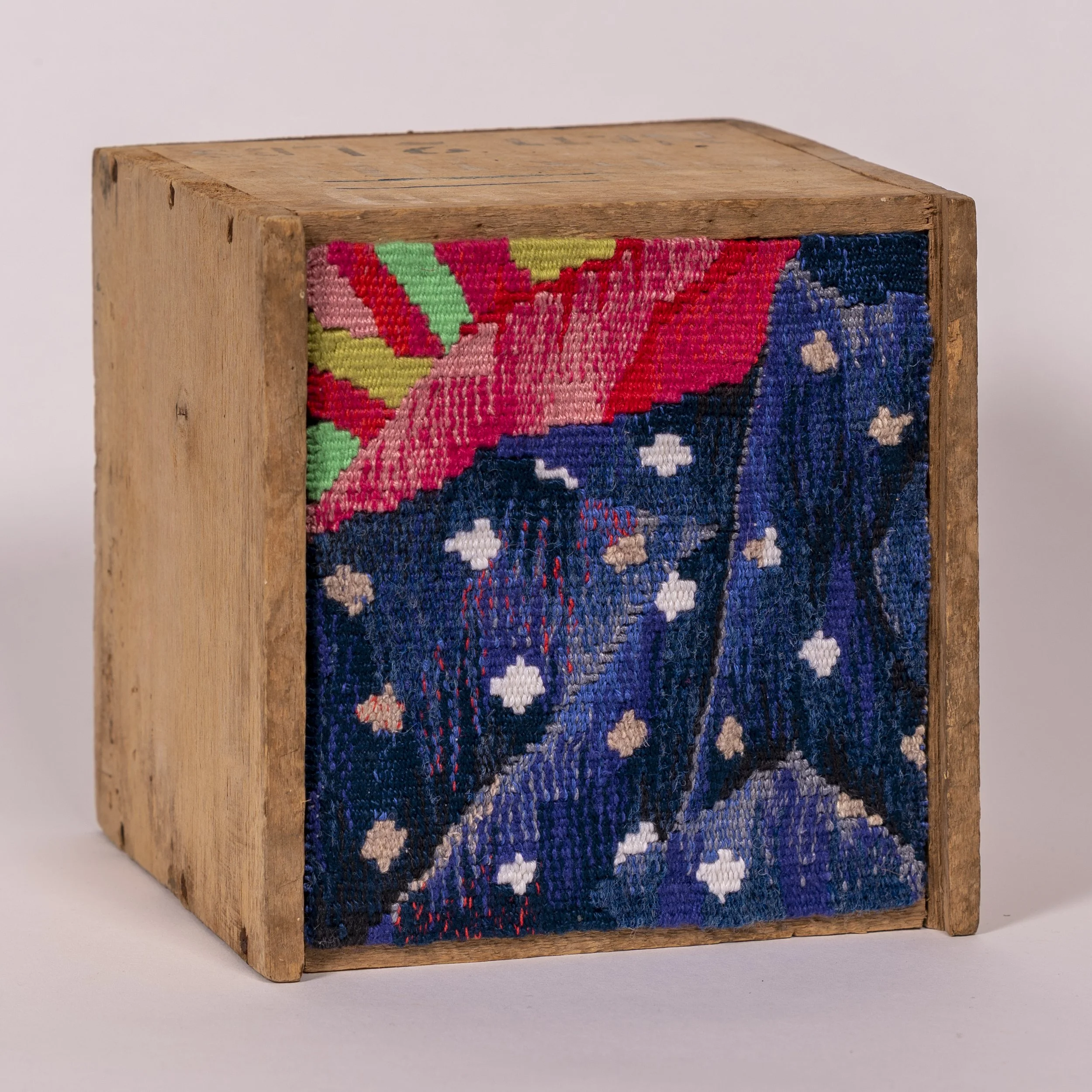 Close-up of a wooden block with colorful woven textile attached to one side, displaying floral and leaf patterns in shades of pink, green, red, blue, black, and white.