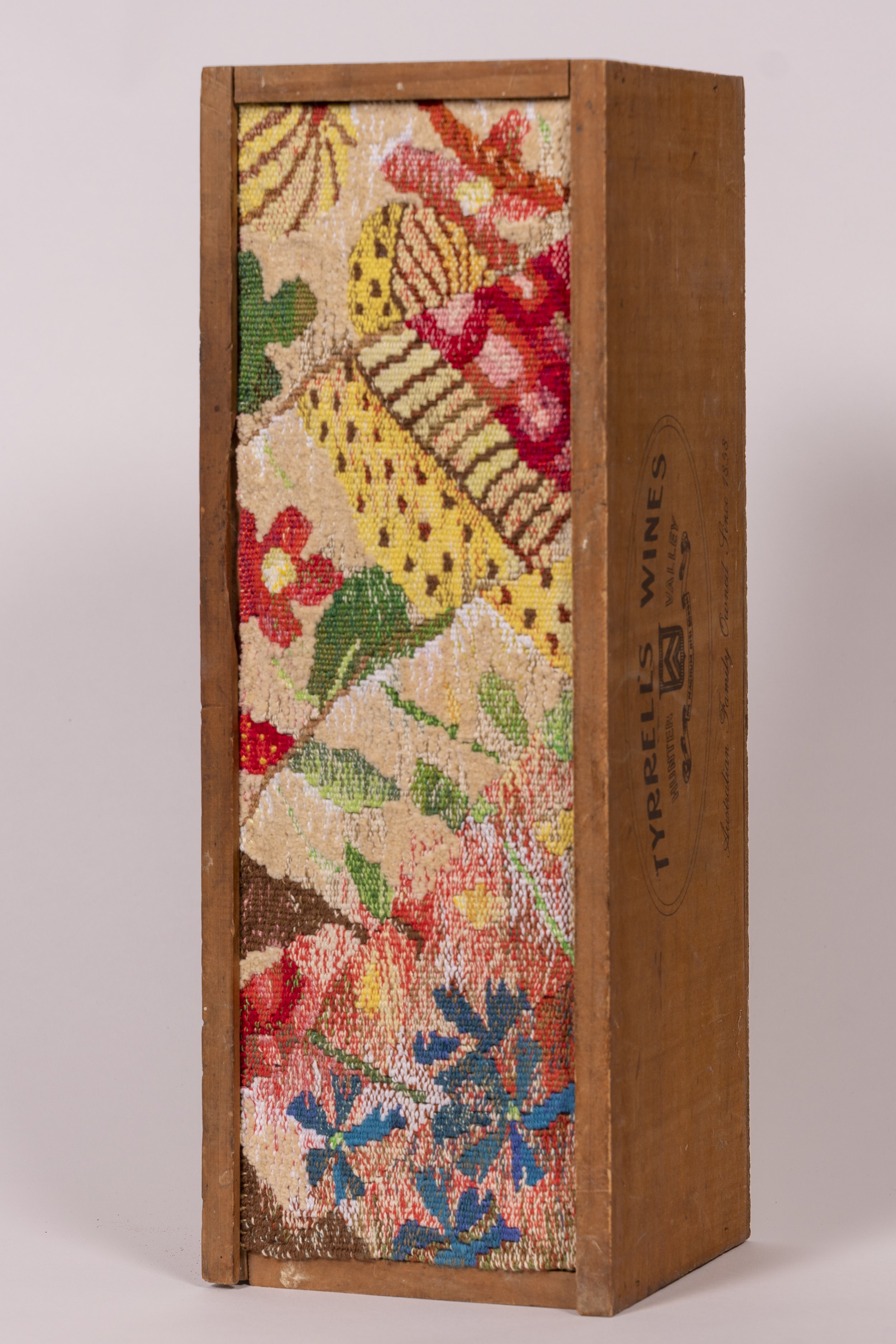 A wooden box with a colorful needlepoint embroidered panel featuring flowers and leaves, set against a plain background.