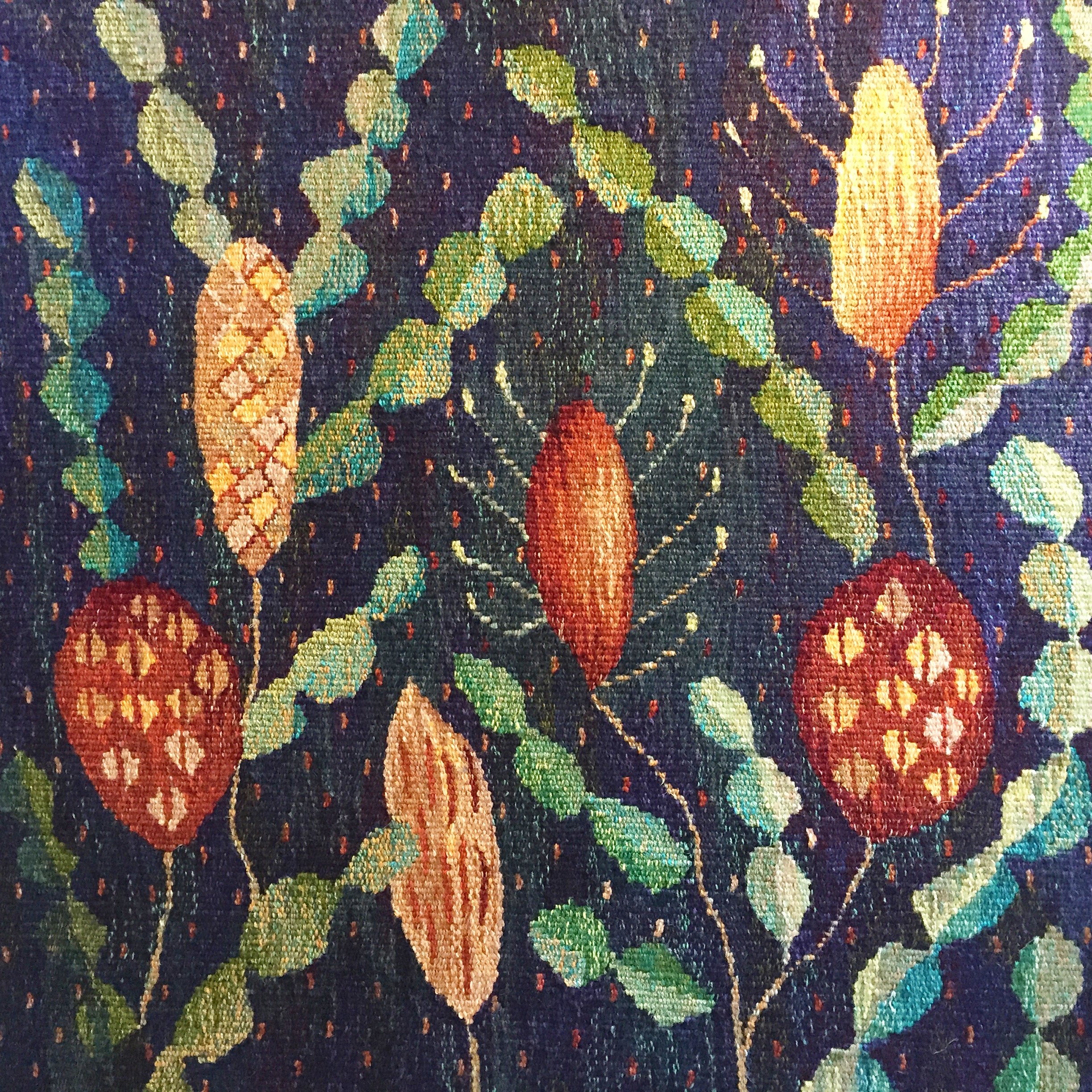 A colourful woven tapestry featuring a pattern of Australian plants with green leaves, orange flowers, and small red dots on a dark background.