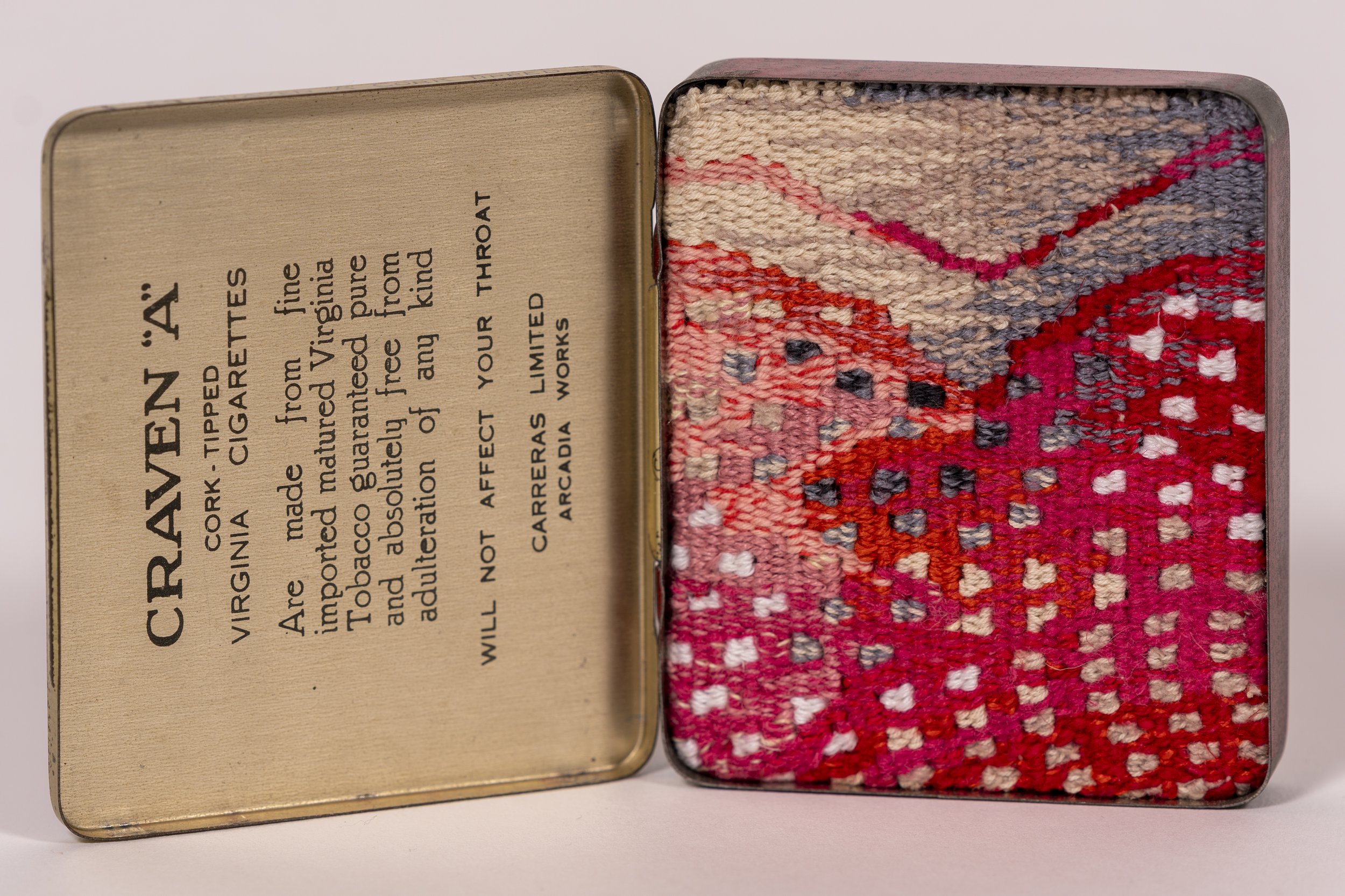 A vintage tin container with a colorful embroidered fabric interior. The tin's lid has printed text describing cigars, including warnings against smoking during pregnancy.