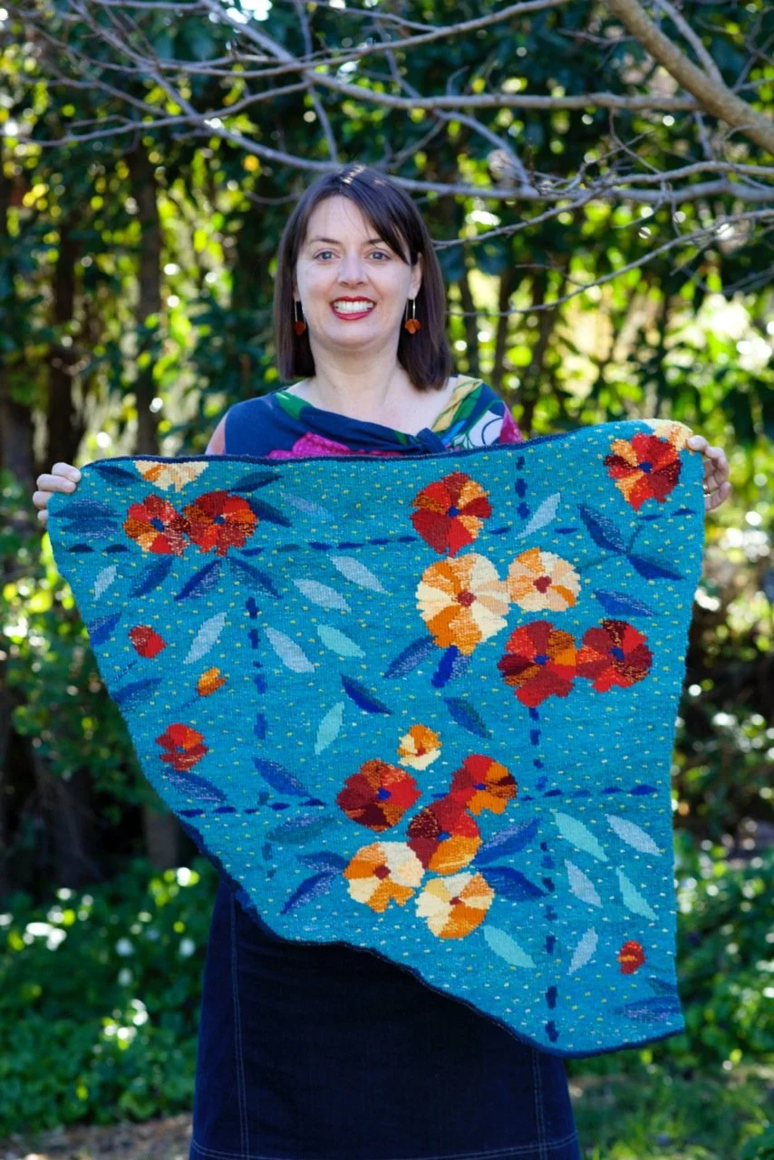 Dimity Kidston holding a colorful woven tapestry based on ann fragment of old lino, outdoors with green foliage background.