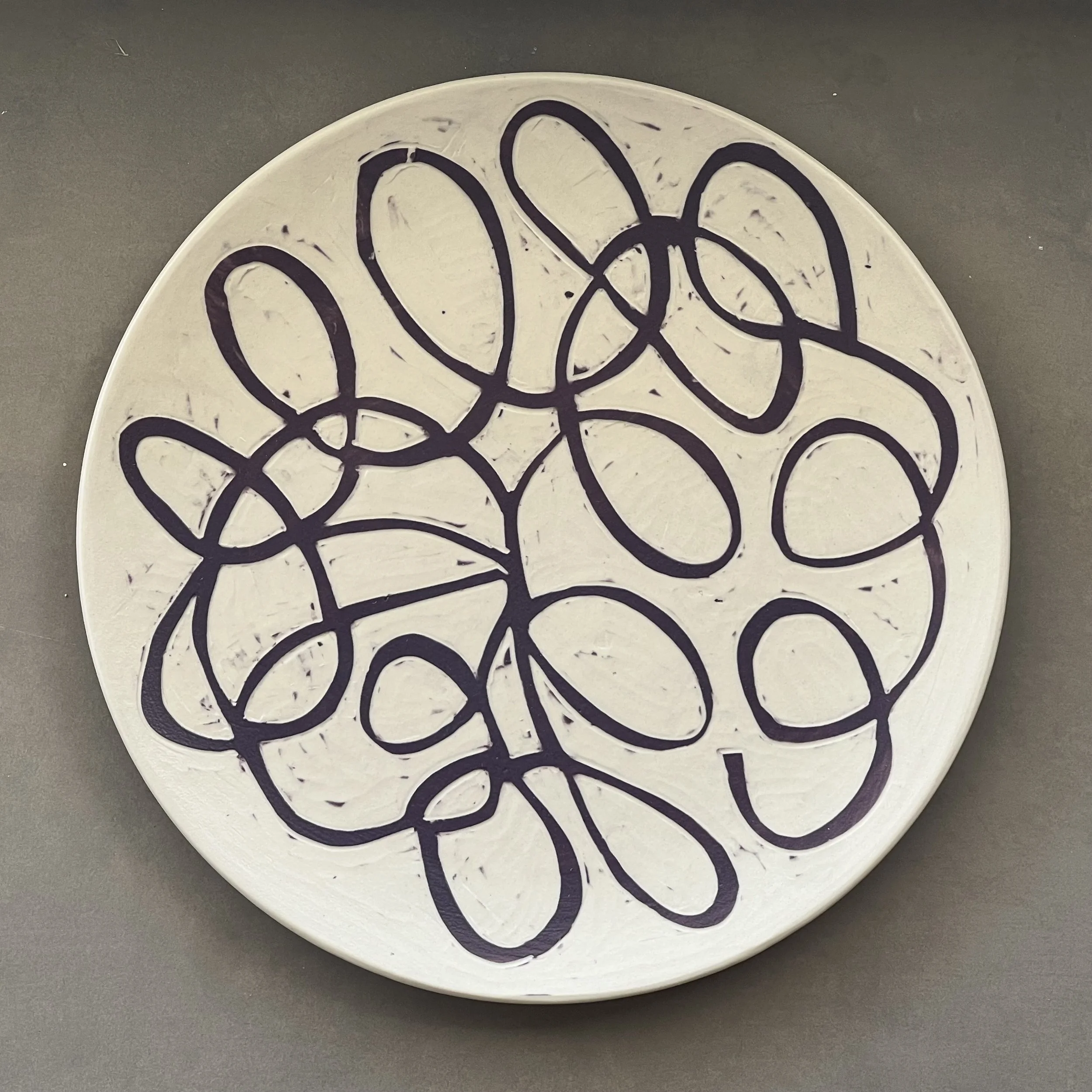 Dimity Kidston sgraffito hand carved ceramic plate with a black abstract line design on a white background, placed on a gray surface.