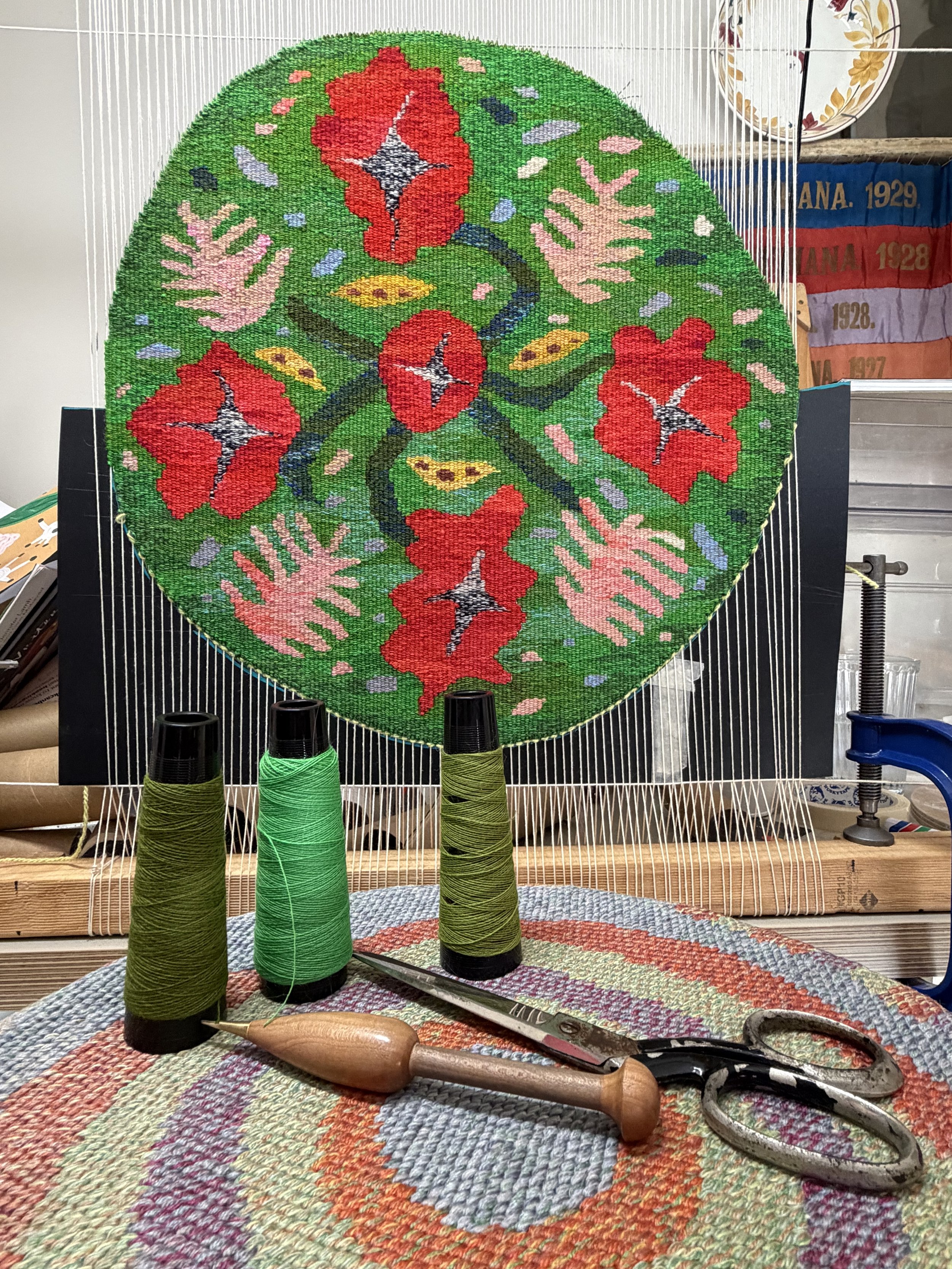 A partially completed, colorful woven tapestry in a circular shape, with various patterns and floral designs, held on a loom with spools of green thread, scissors, and a crochet hook in the foreground.