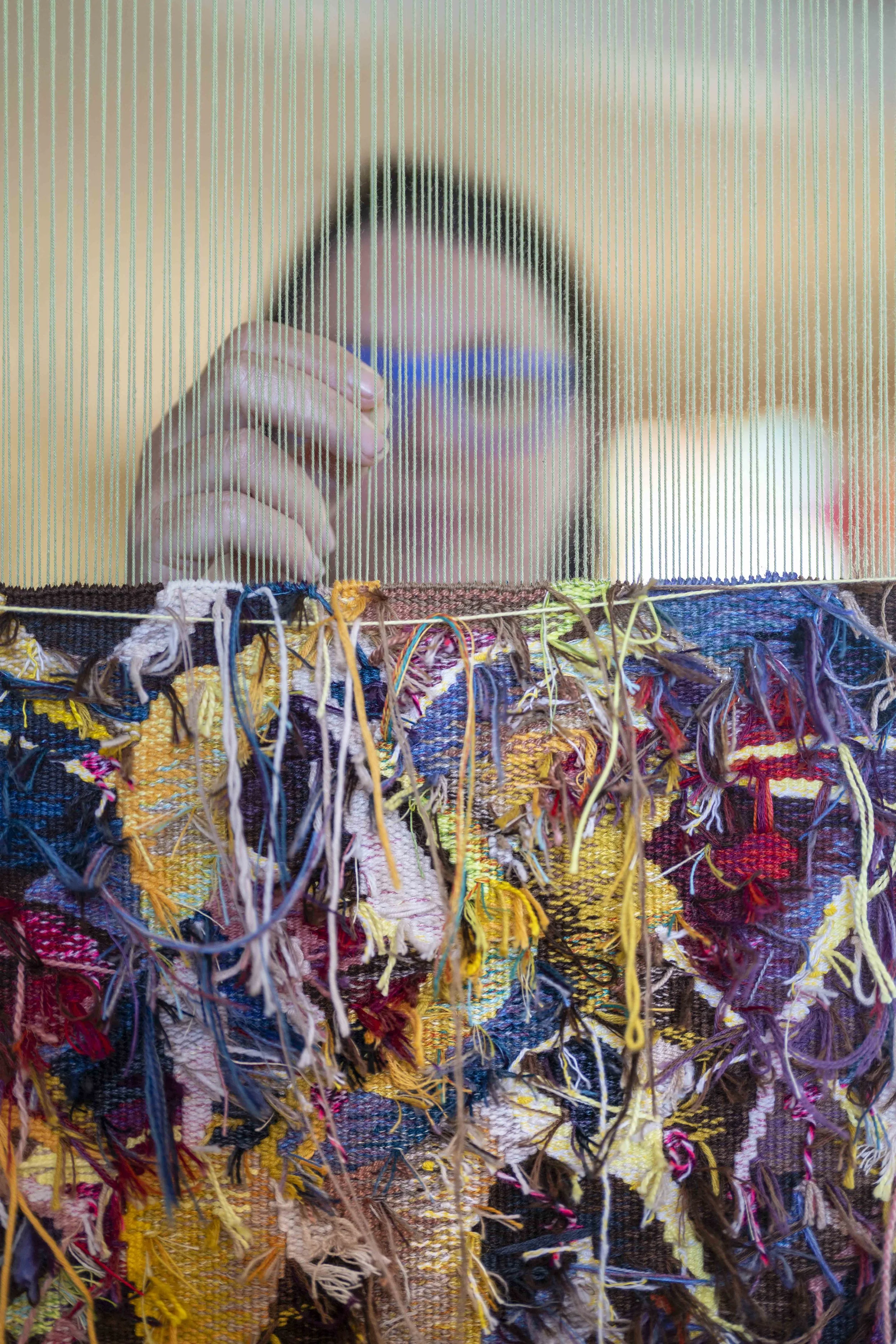 Close-up of Dimity Kidston weaving a colorful tapesrty  on a loom, with her hand pulling threads through the woven textile.